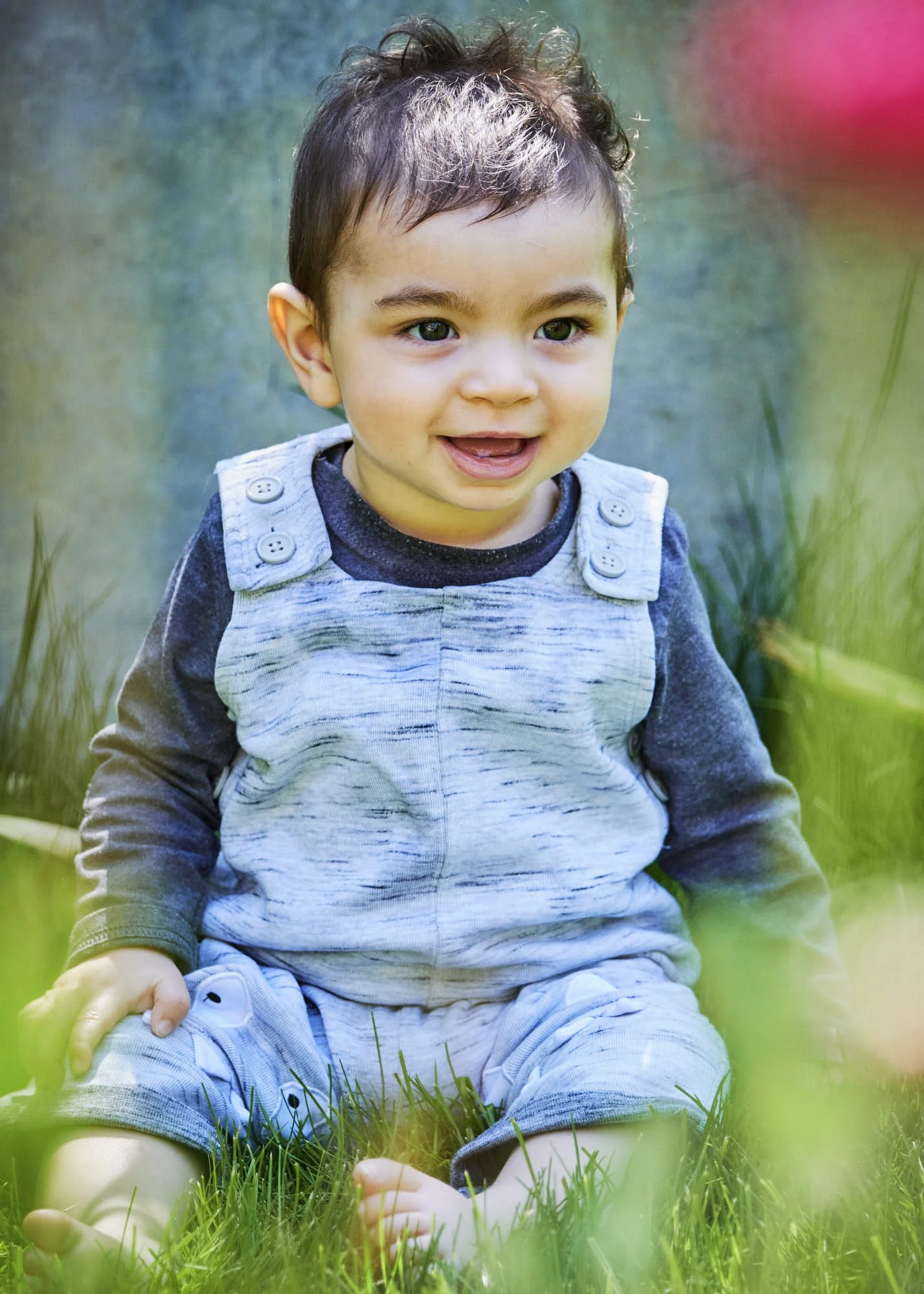 A young boy with curly brown hair, sitting on the grass outdoors, wearing a blue and gray long sleeve shirt with buttons on the shoulders, smiling and looking to the side.