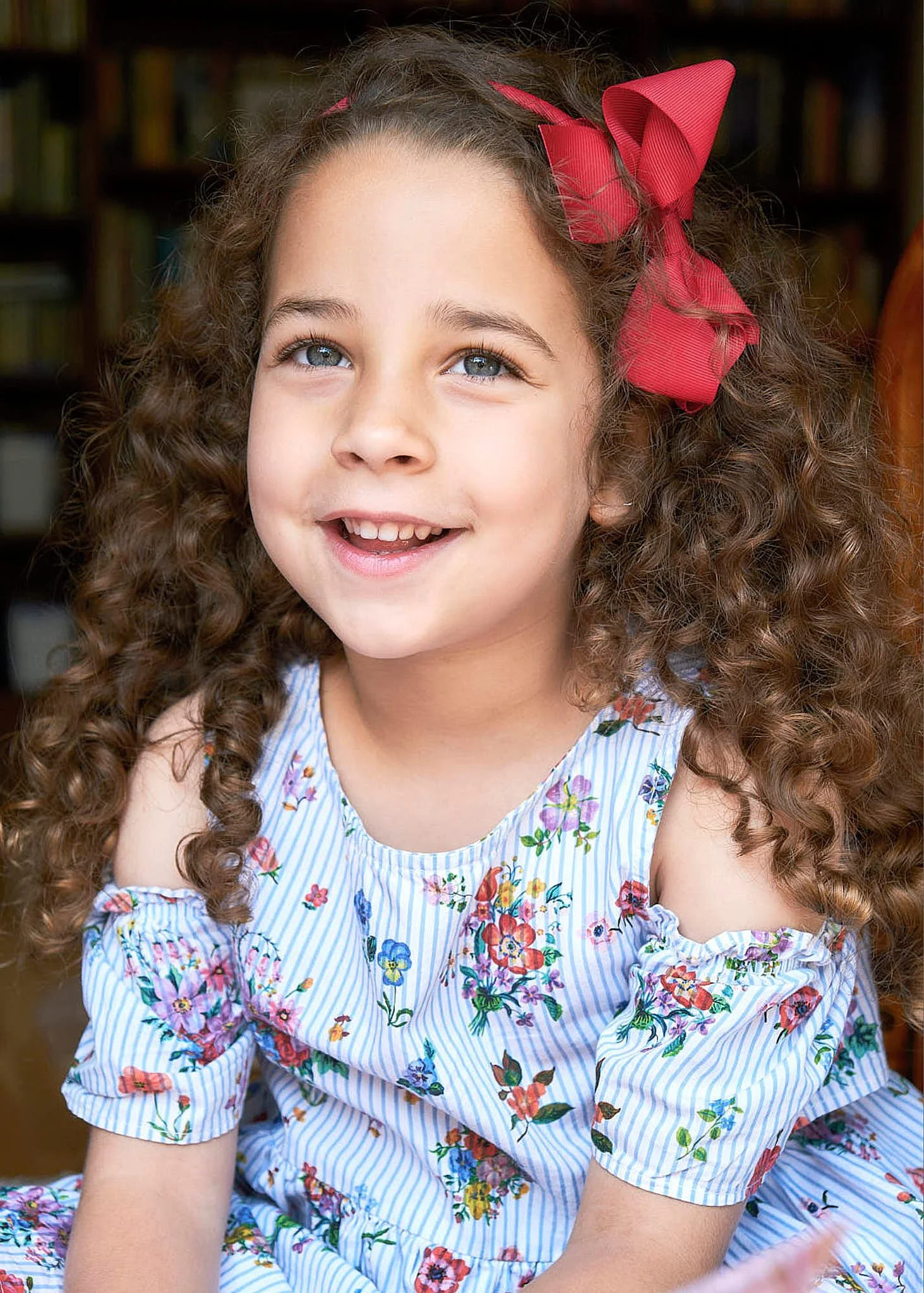 A young girl with curly brown hair and blue eyes, wearing a blue and white striped dress with colorful floral patterns, smiling and looking slightly upward, with a large red bow in her hair.