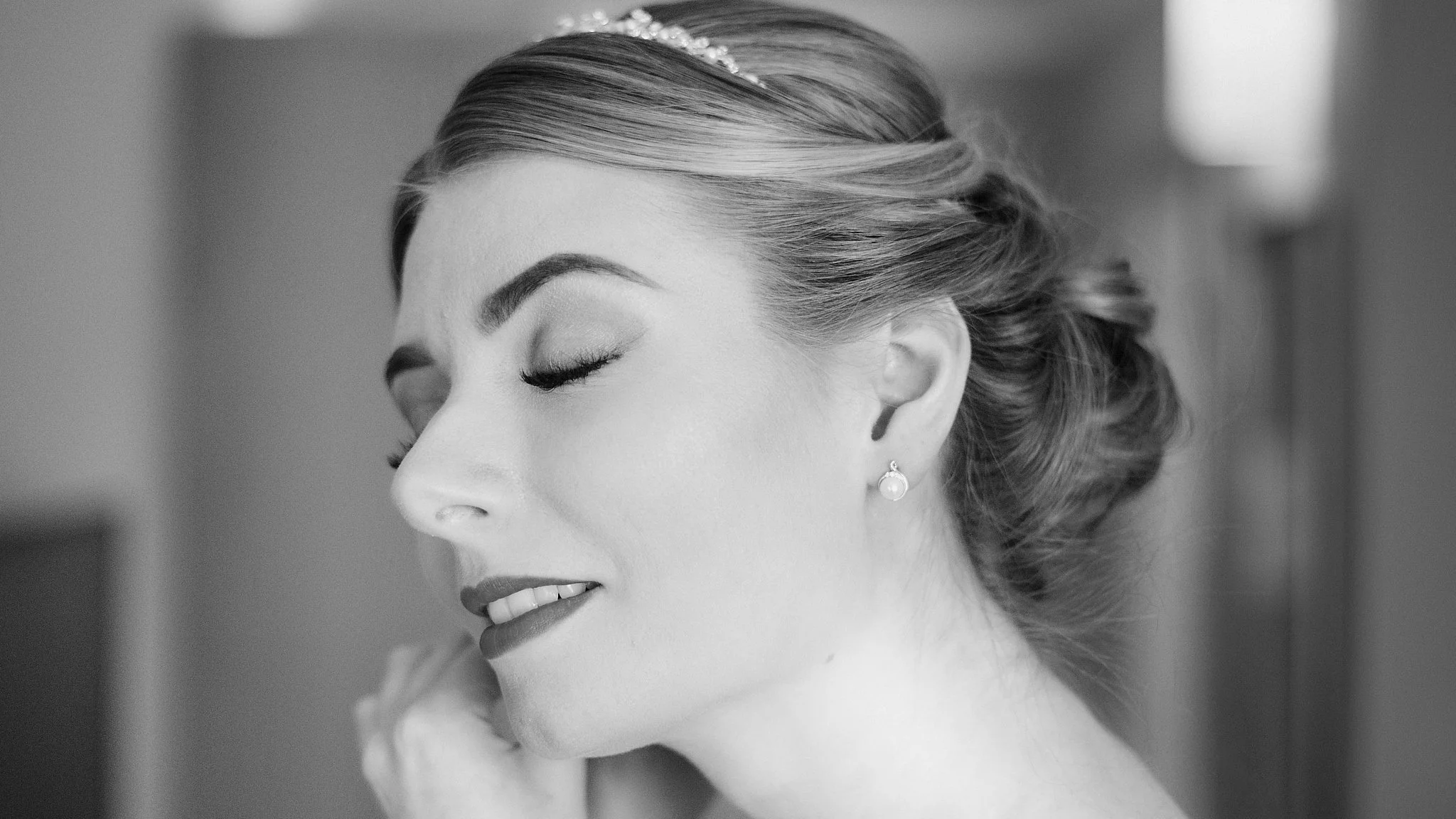 A close-up black and white portrait of a woman with her eyes closed, wearing pearl earrings and a headband with decorative details, with her hair styled in an elegant updo.