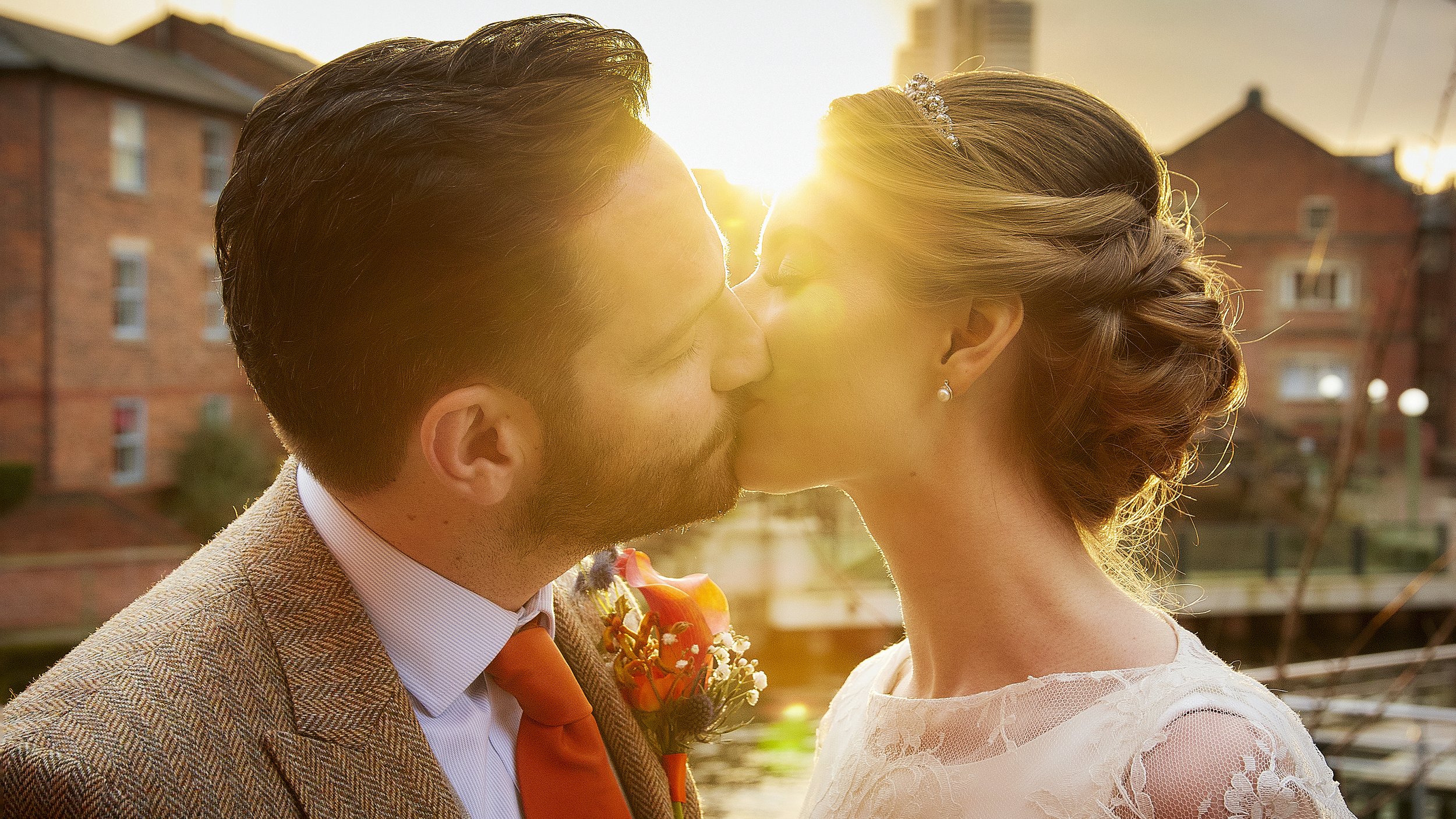 A bride and groom sharing a kiss outdoors during sunset, with the sun shining behind them and a brick building in the background.
