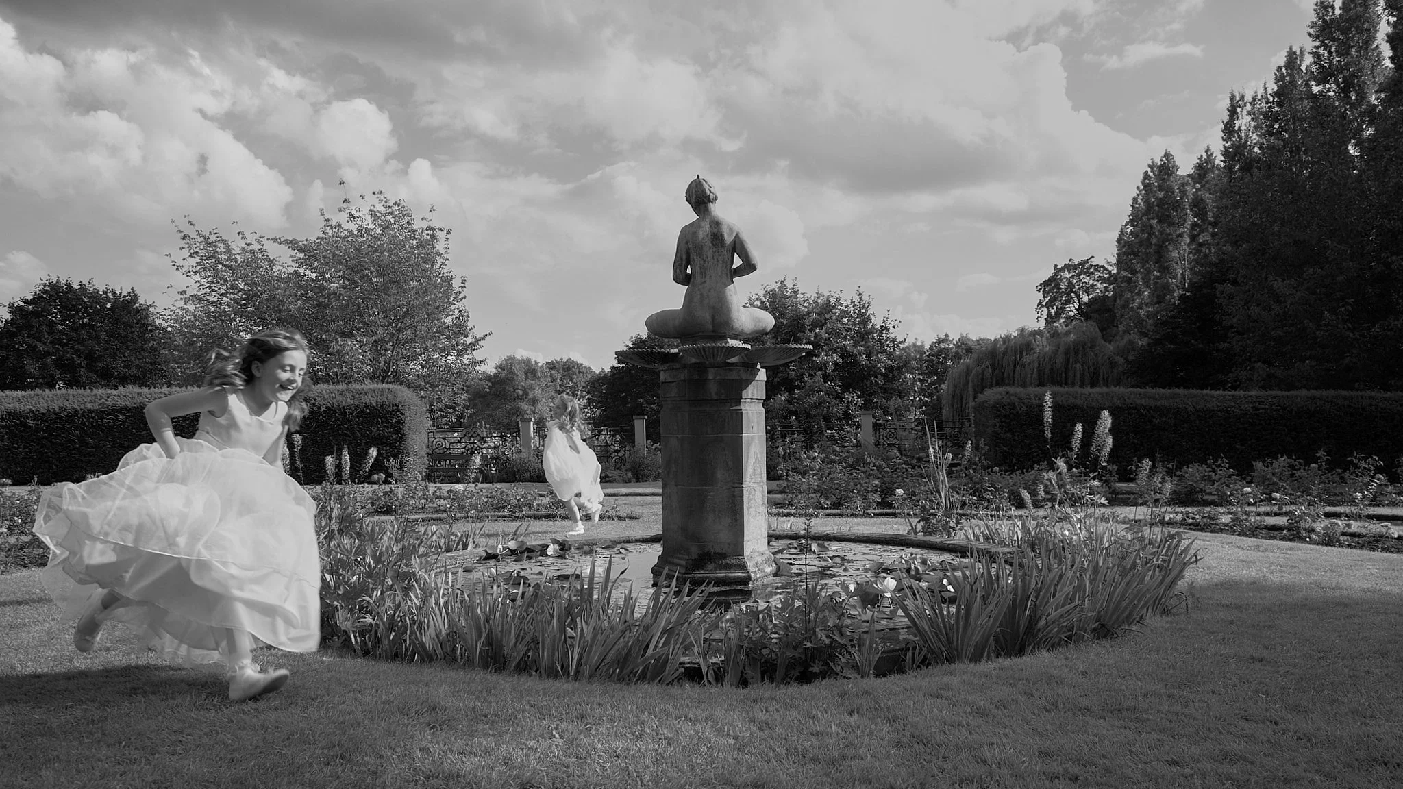 Two young girls in dresses playing and running outside near a fountain with a statue of a seated person, surrounded by greenery and trees, under a partly cloudy sky.