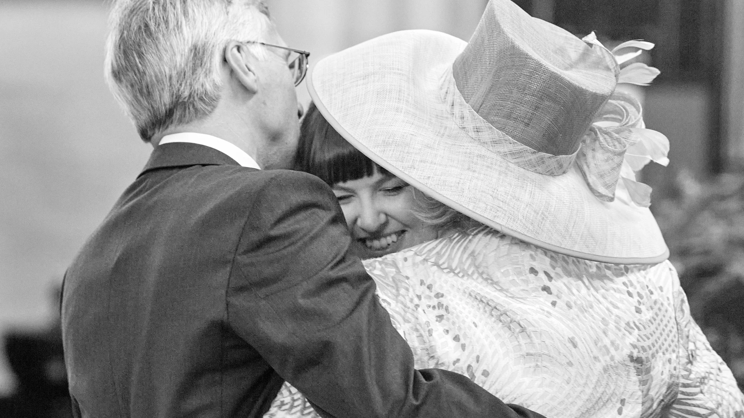 A black and white photo of a man in a tuxedo embracing a woman wearing a large, decorative hat and a patterned dress, both smiling with eyes closed.