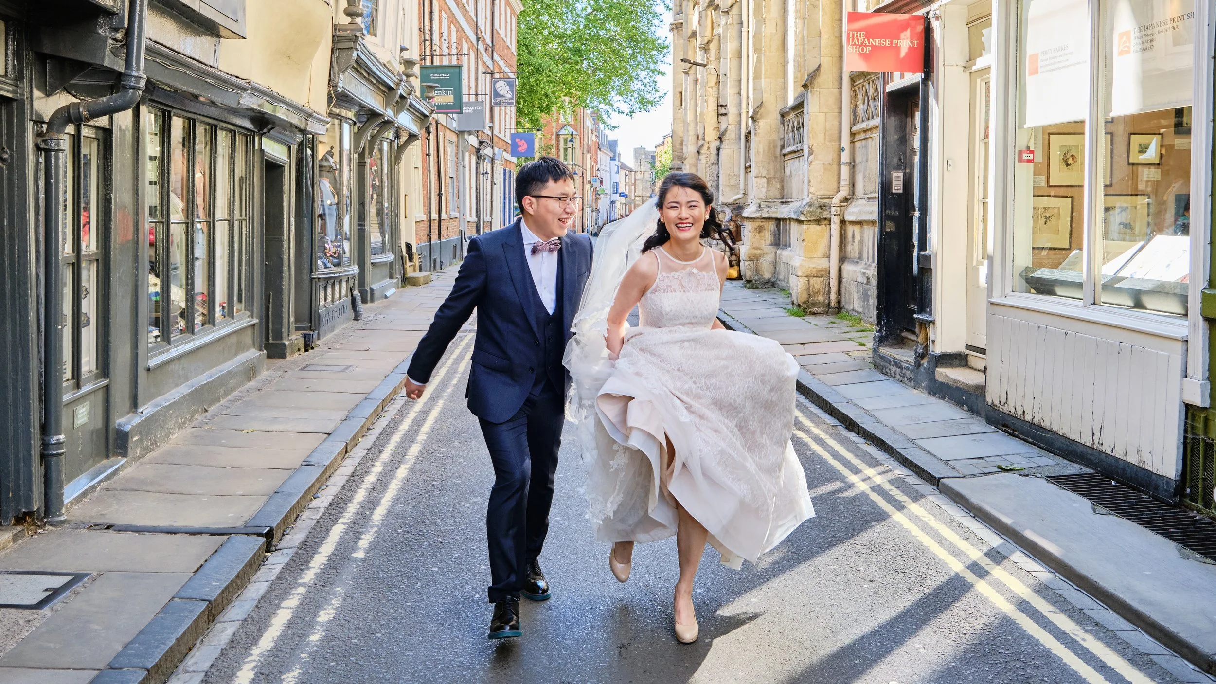 A newlywed couple walking and laughing on a city street, with the bride lifting her wedding dress and the groom in a suit and bow tie.