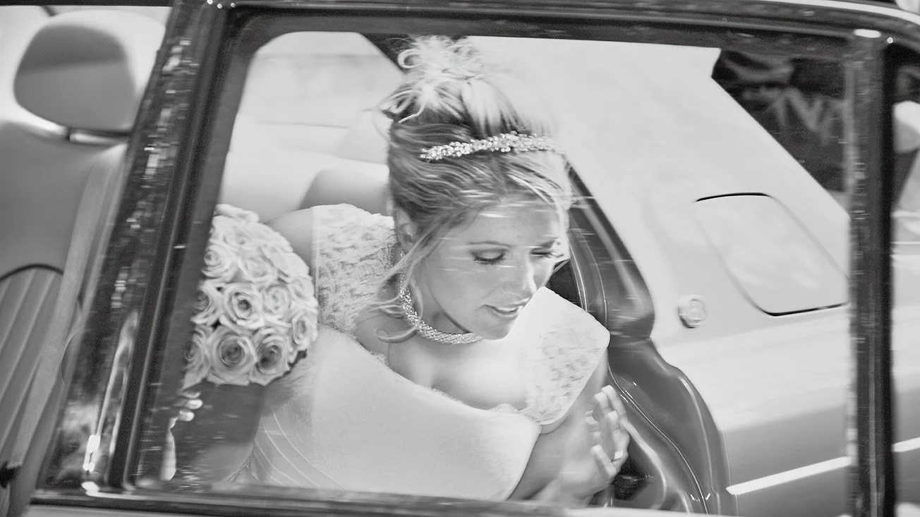 A bride in a wedding dress with a bouquet of roses and a tiara on her head, sitting in a car, looking down with her hands pressed together.