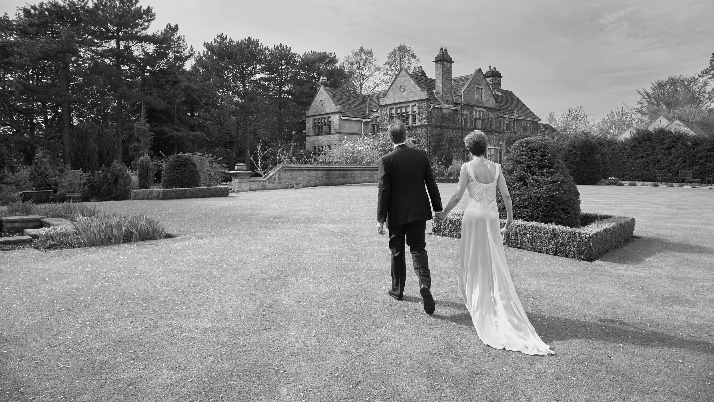 A couple dressed in wedding attire walking hand in hand in a garden with a large historic stone mansion in the background.
