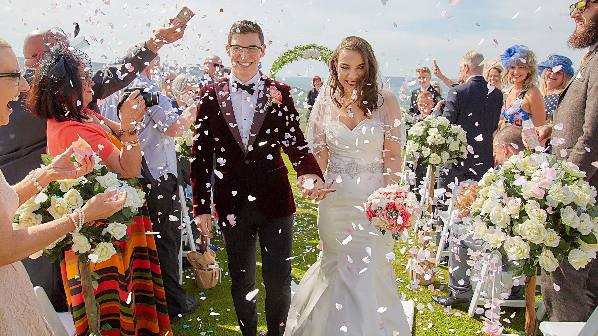 A bride and groom walking hand in hand down the aisle at their outdoor wedding ceremony, surrounded by friends and family with flower arrangements and confetti falling.