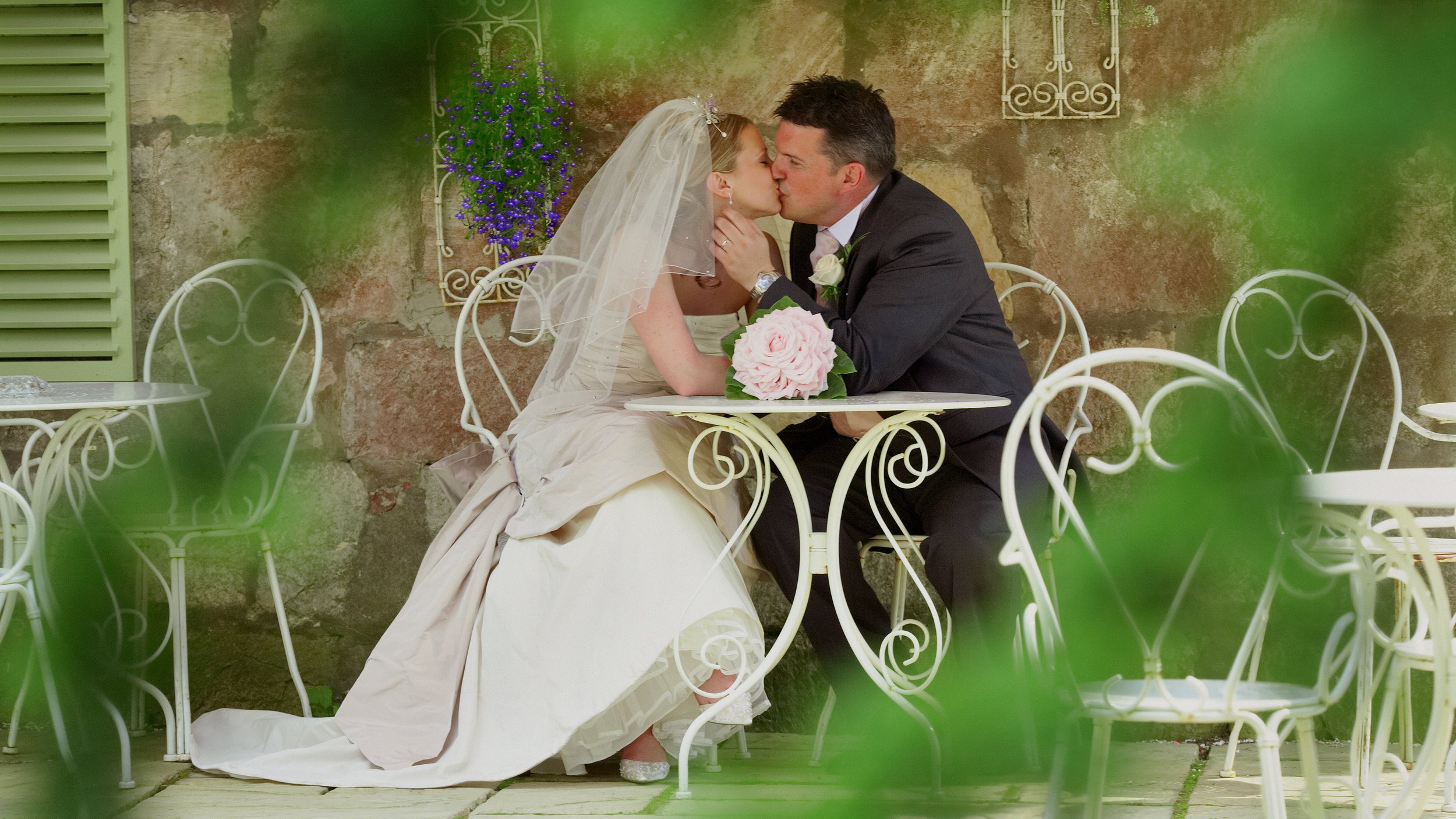 A bride and groom share a kiss at an outdoor wedding reception, surrounded by white wrought iron chairs and a rustic background with greenery in the foreground.