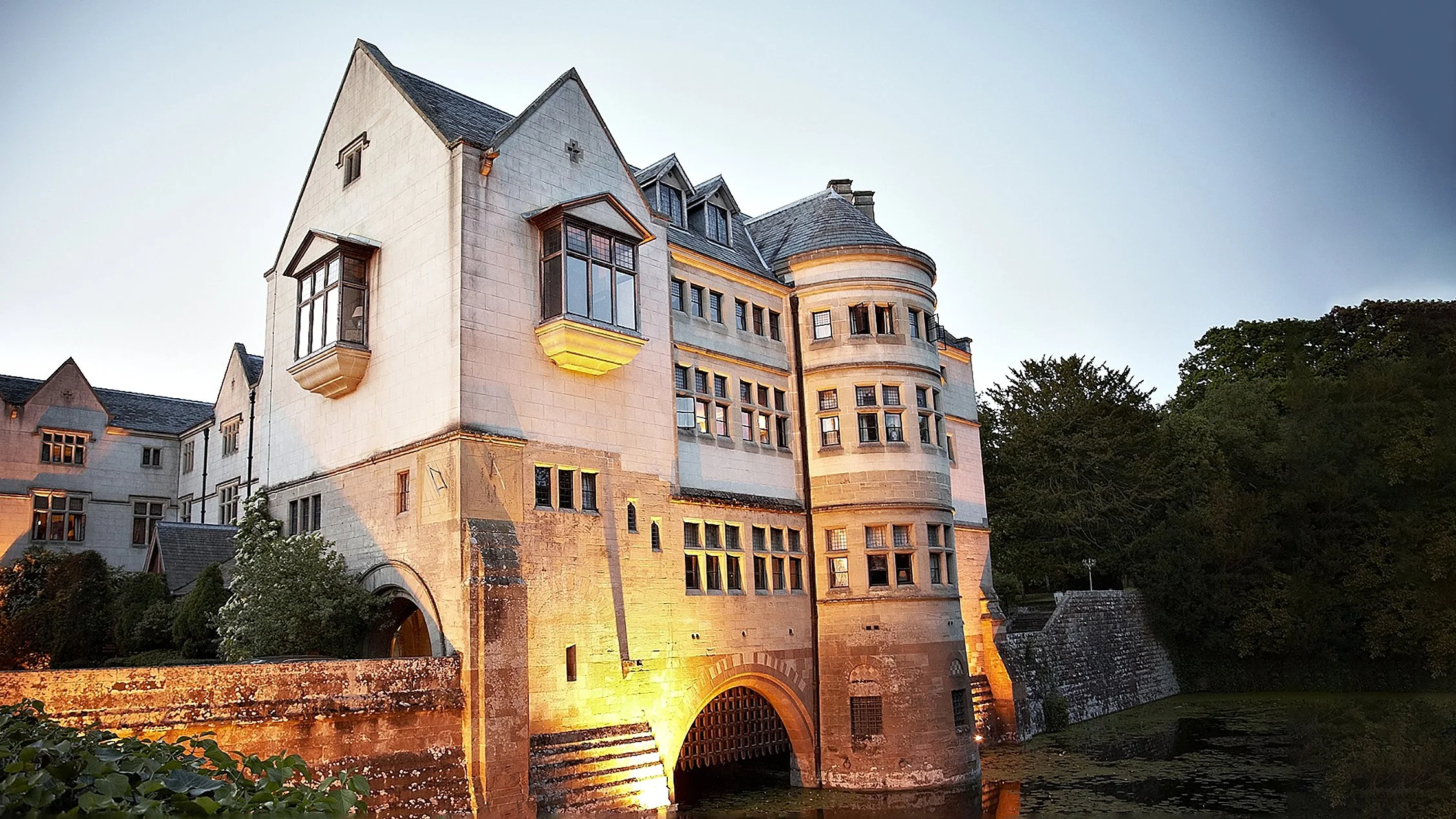 A large historic castle-like building with multiple turrets, windows, and brick arches over a waterway, illuminated at dusk.