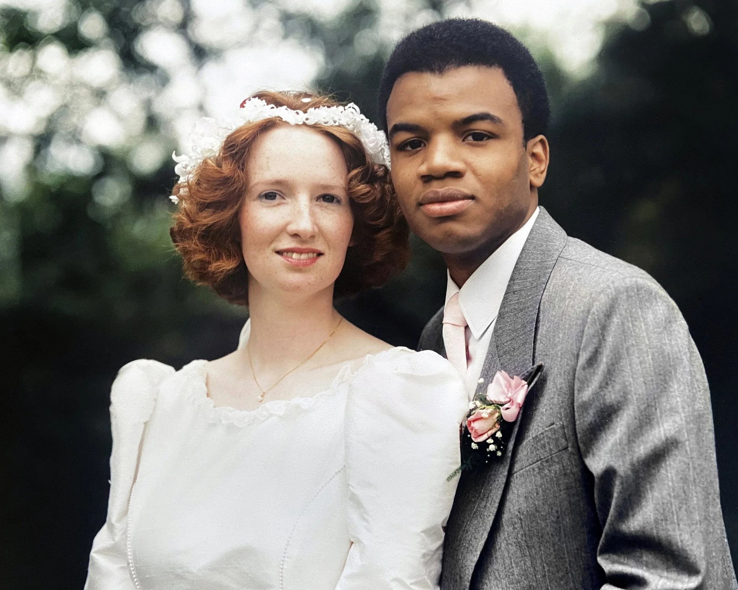 A newlywed couple posing outdoors, the woman with red hair and a lace veil smiling, and the man with short dark hair wearing a grey suit with a pink tie and boutonnière, both looking at the camera against a blurred green leafy background.