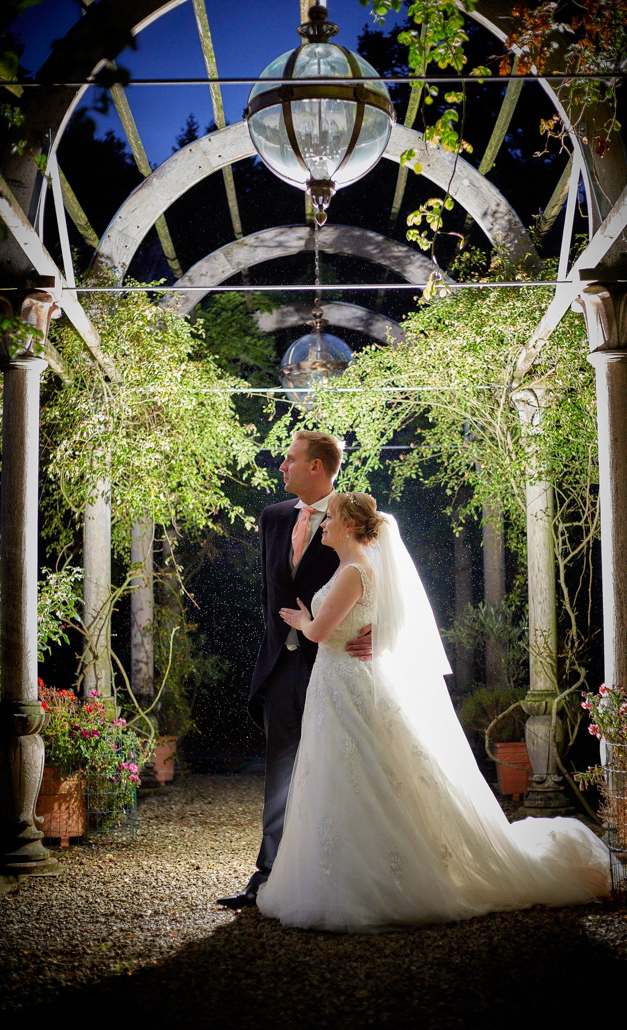 A bride and groom are standing under a decorative archway at night, surrounded by plants and flowers, with a lantern hanging above them. The bride is wearing a white wedding dress with lace details, and the groom is in a black suit with a pink tie. T