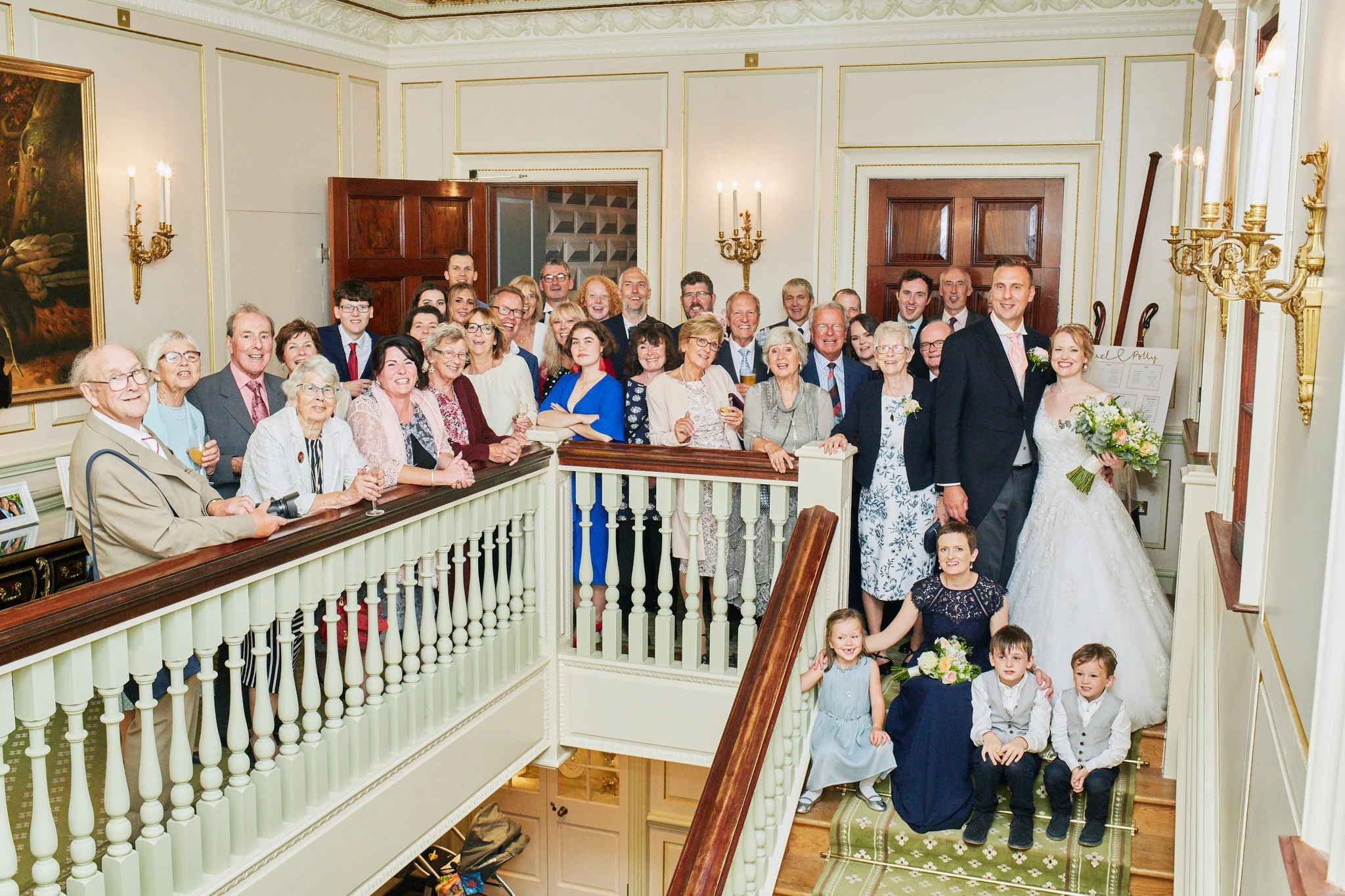 A large group of people, including children, gathered on a staircase and landing inside a decorated elegant room for a wedding celebration. The bride and groom are at the front on the right, smiling, with the bride holding a bouquet. The group is smi