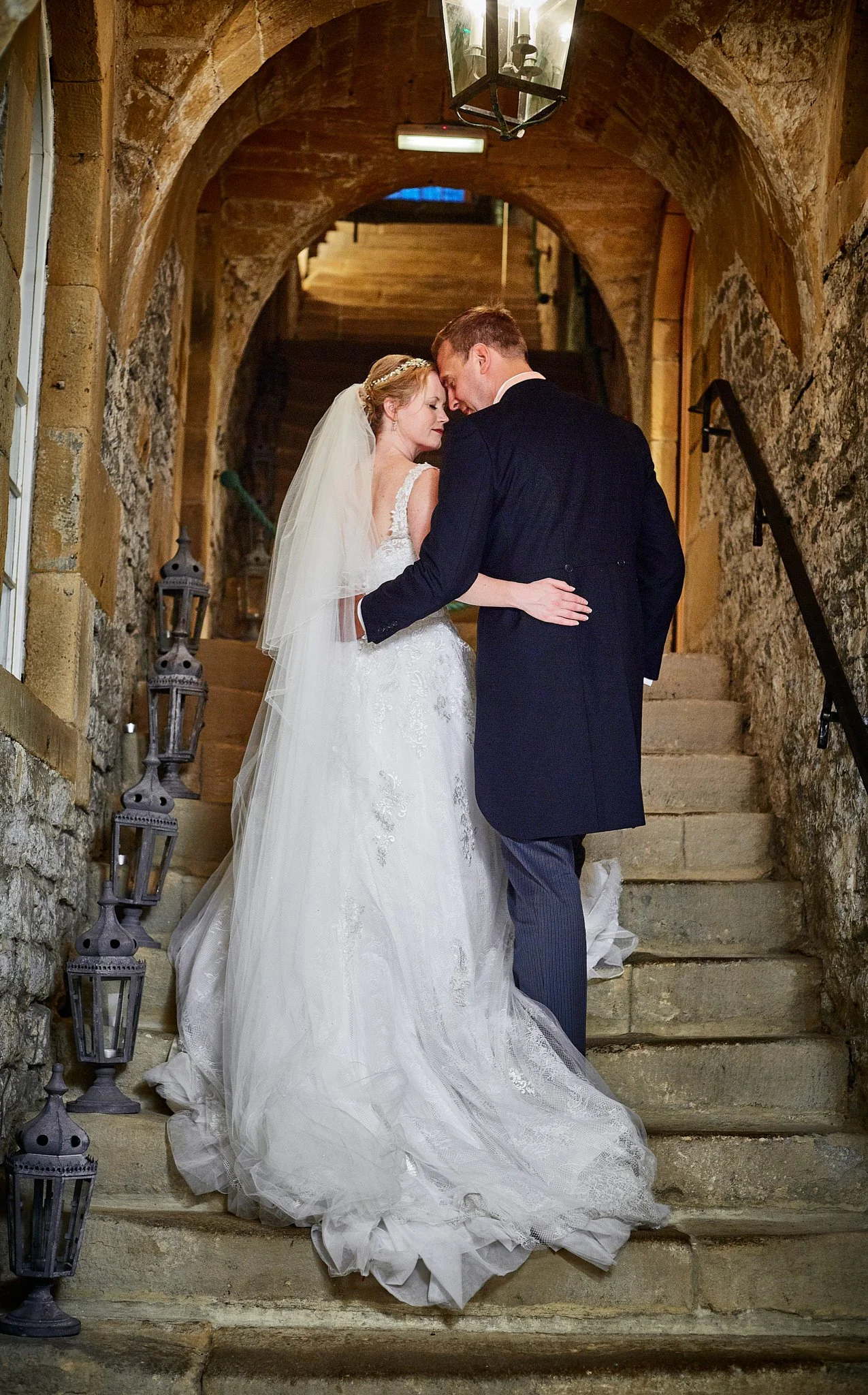 A bride and groom sharing a tender moment on stone staircase in a rustic setting, with lanterns decorating the walls.