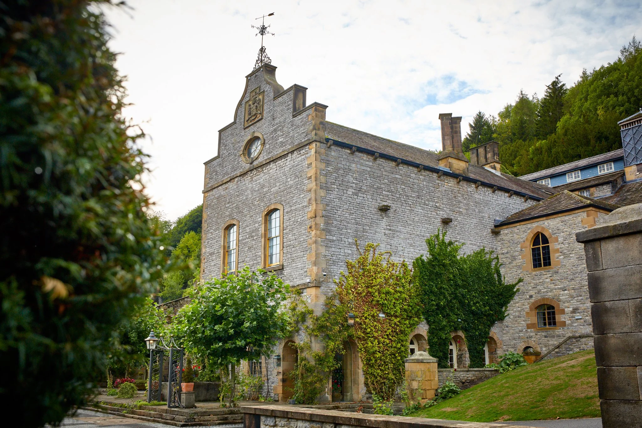 A stone church with a clock and a coat of arms on its front facade, surrounded by greenery, with trees in the background and a partly cloudy sky.