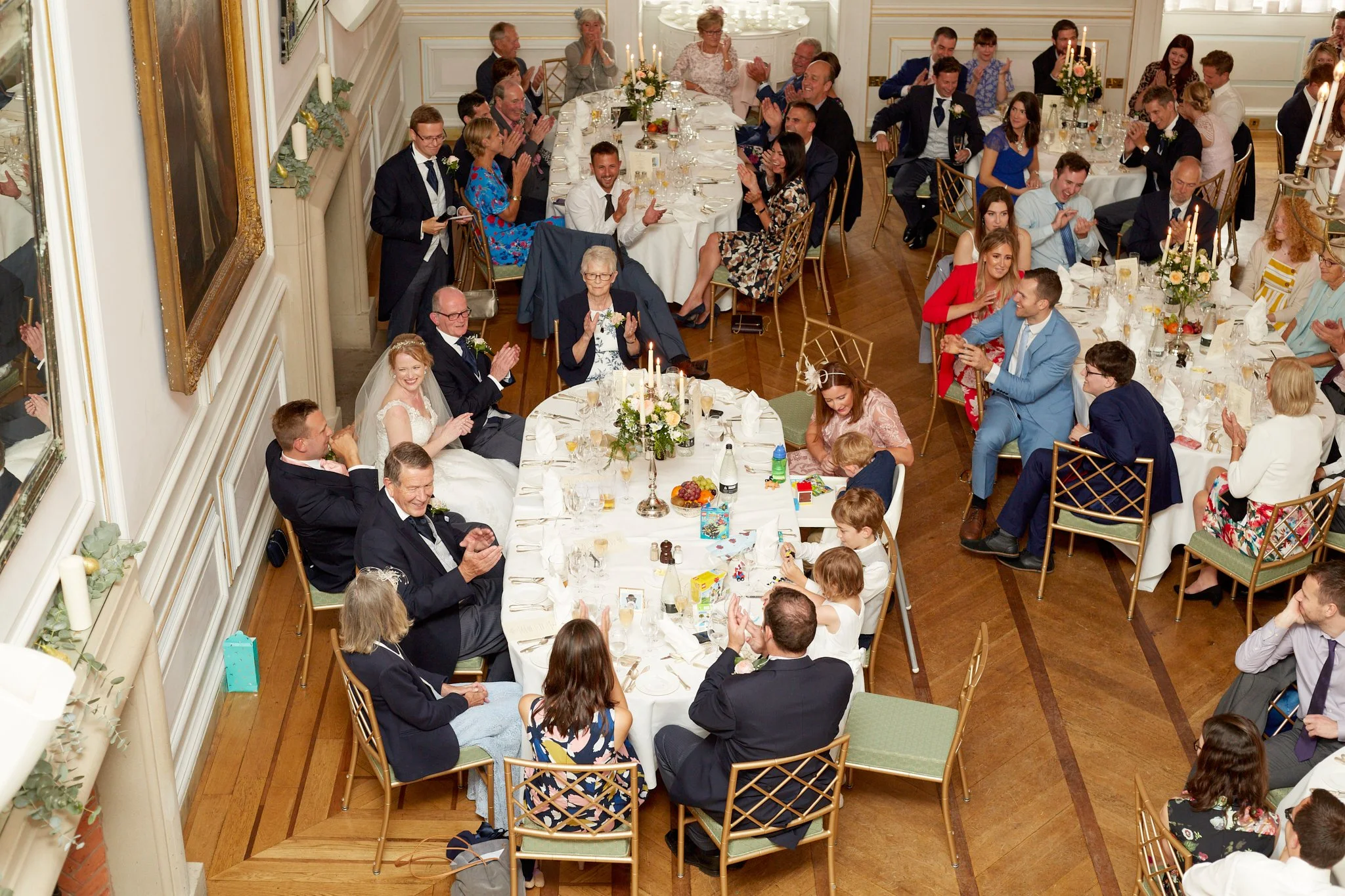 A large group of people gathered at round tables, celebrating a wedding reception in an elegant room with white walls, decorated with candles and floral arrangements. The bride and groom are seated at one of the tables, smiling and surrounded by frie