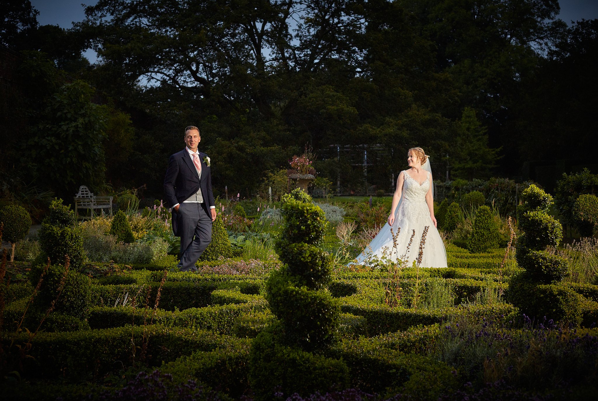 A bride and groom standing in a garden at dusk. The groom is wearing a dark suit with a pink tie and boutonniere. The bride is in a white wedding gown with lace details, looking at the groom with a smile. The garden is lush with green bushes and flow