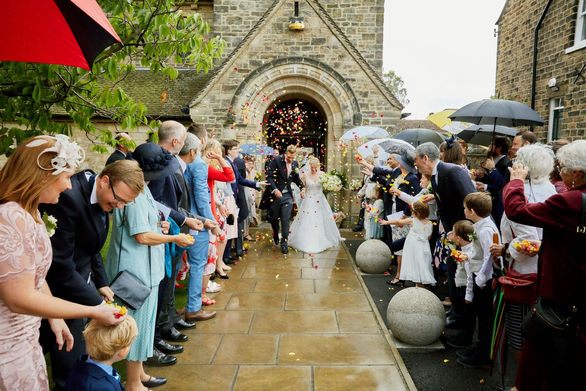 A diverse group of people celebrating a wedding outside a stone church, with confetti and umbrellas. The bride and groom are walking hand in hand, smiling, as guests throw flower petals and celebrate.