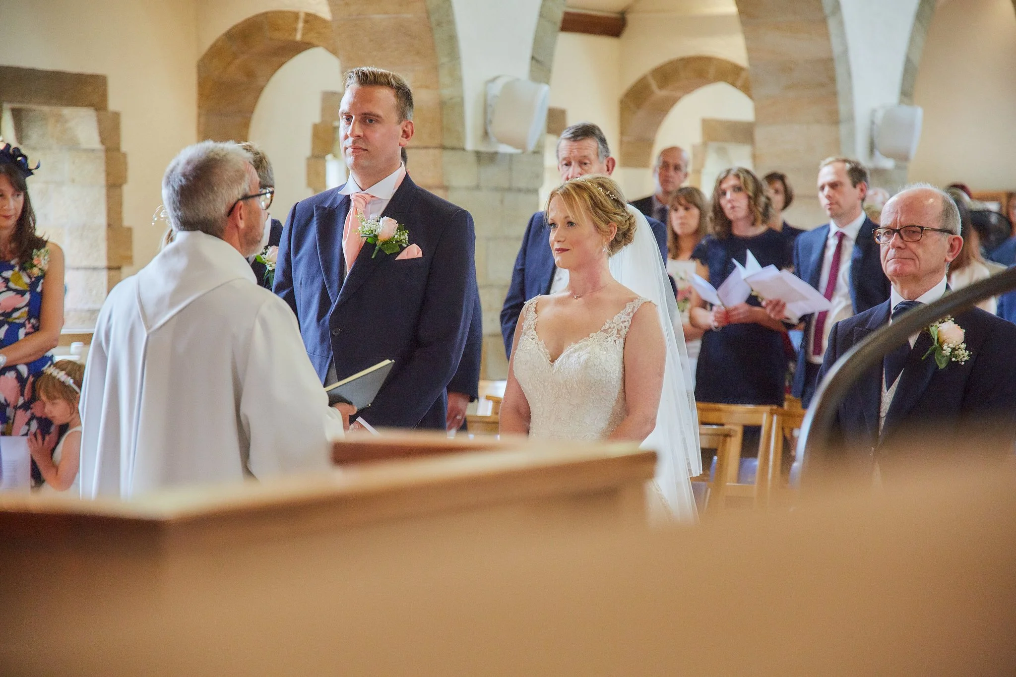 A bride and groom standing before a priest during a wedding ceremony inside a church, with guests seated in the background.