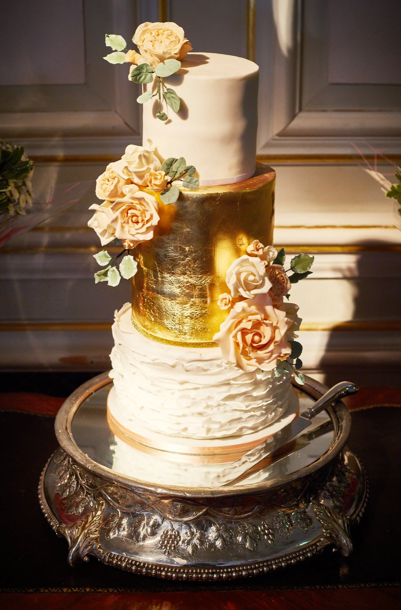 A three-tiered wedding cake decorated with cream, gold, and peach-colored roses and green leaves, placed on an ornate silver cake stand.