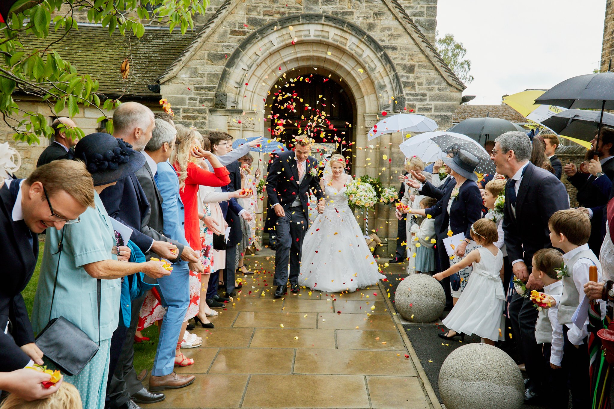 A bride and groom walking out of a church, surrounded by guests throwing flower petals, with umbrellas and formal attire on a rainy day.