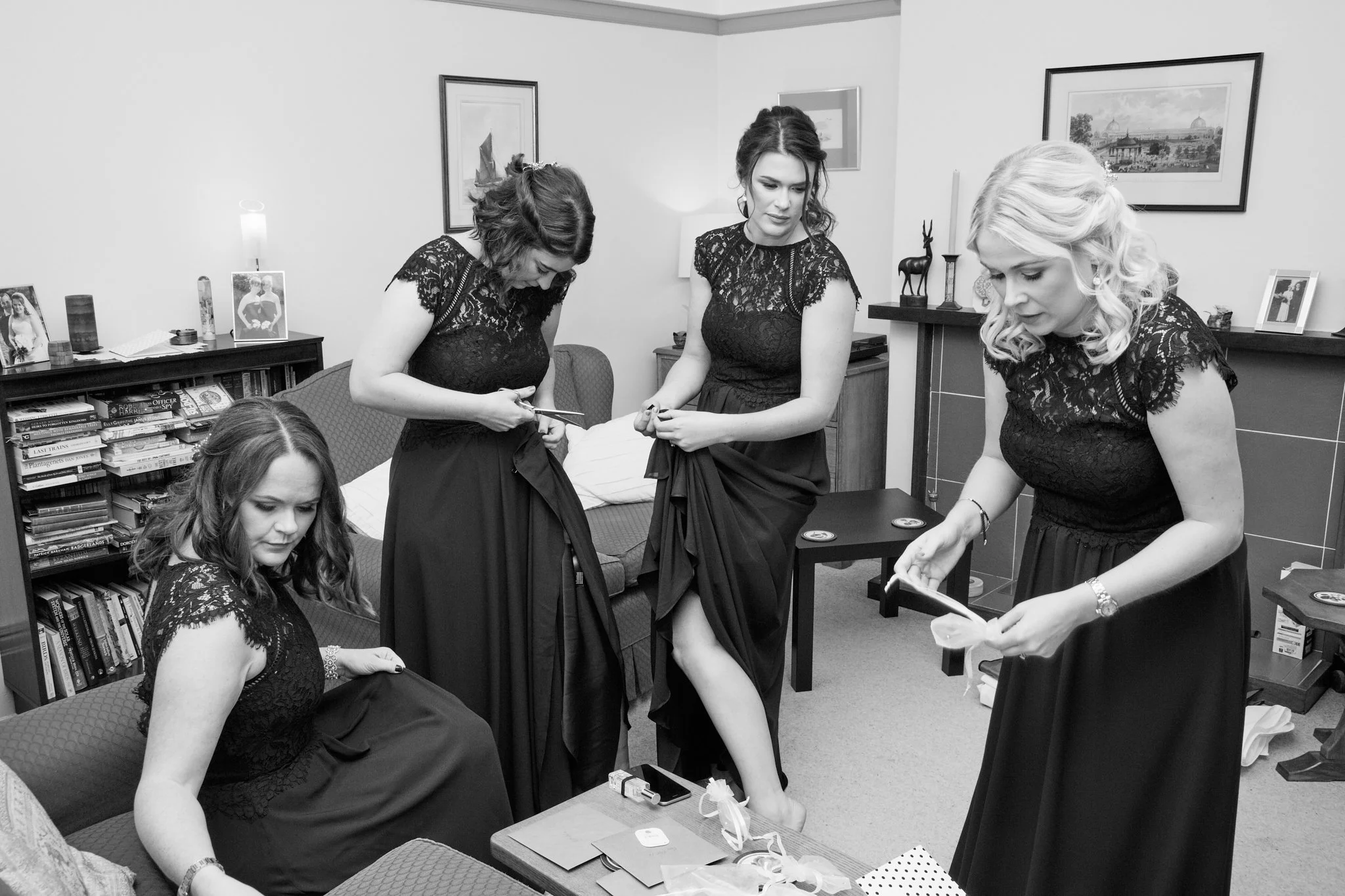 Five women in black lace dresses preparing for a formal event in a living room with framed pictures and books, some sitting on a sofa and some standing, surrounded by various items including a smartphone and gift bags.
