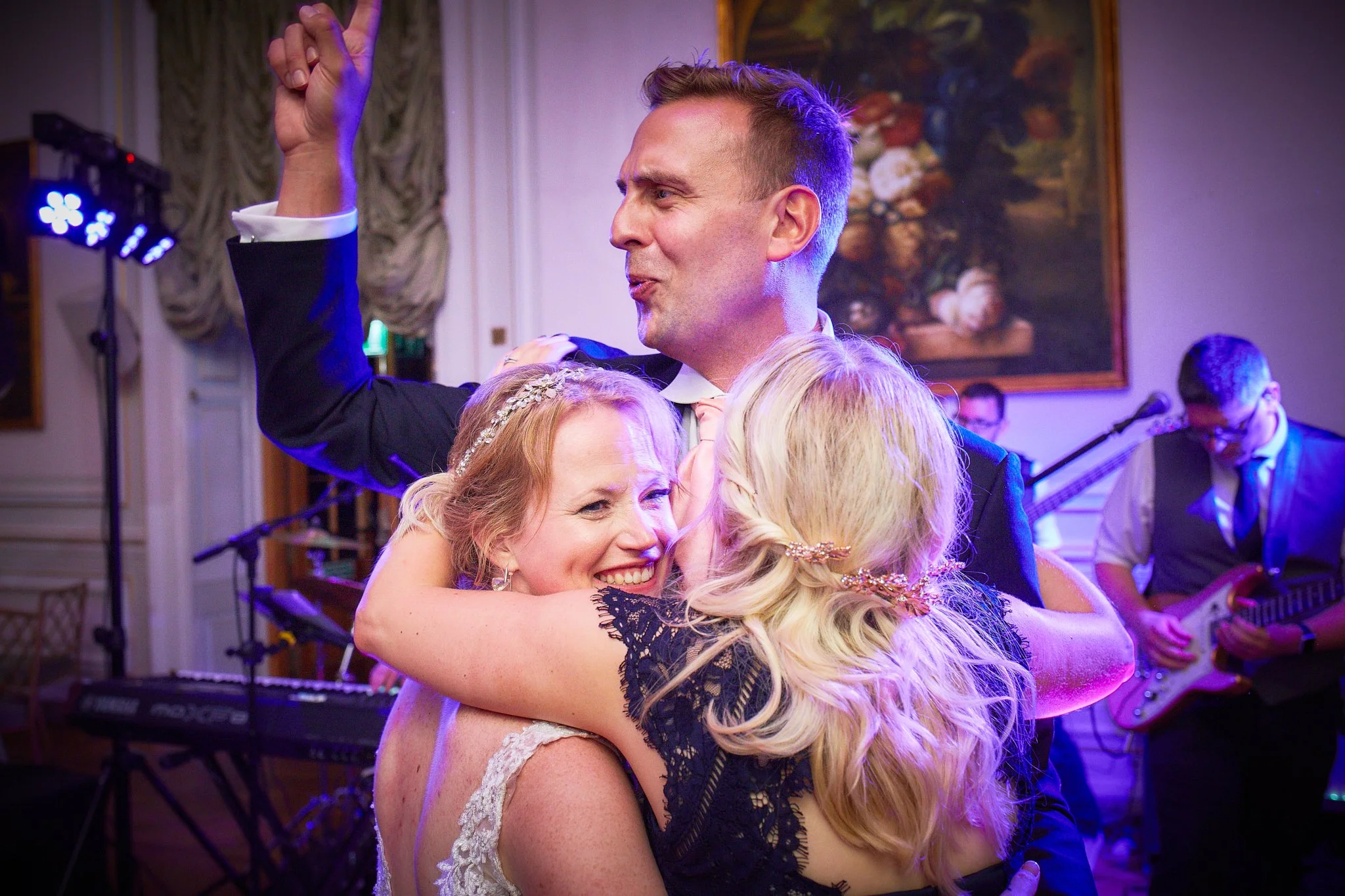 People hugging and smiling at a celebration or wedding reception with a band playing in the background.