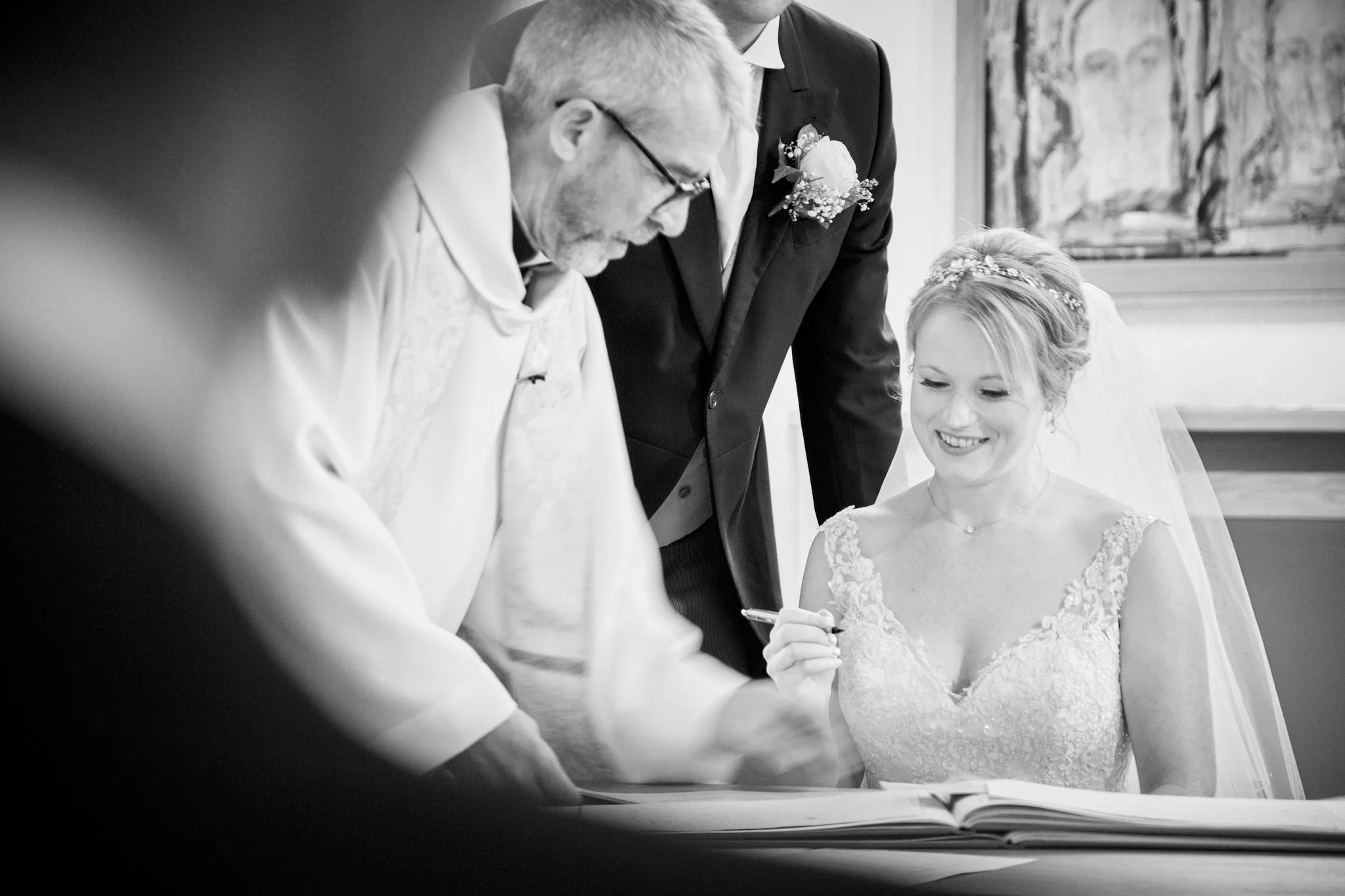 A wedding scene with a bride and two men, all four are signing what appears to be a marriage document. The bride is smiling and wearing a lace wedding gown with a floral headpiece. One man is in a white dress shirt, and the other in a dark suit with 