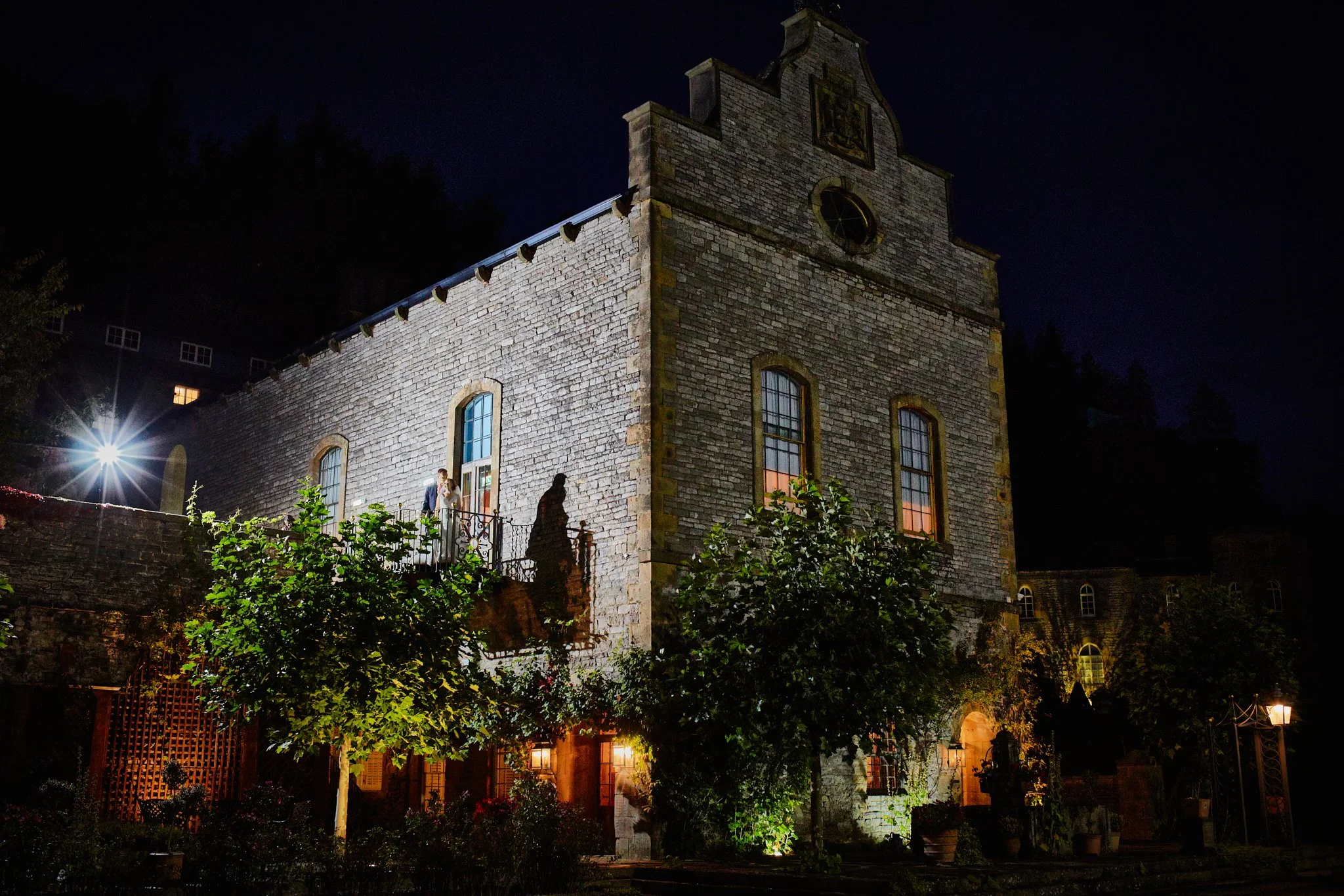 Nighttime view of a historic stone church with illuminated windows and a clock at the front, surrounded by greenery and garden lights.