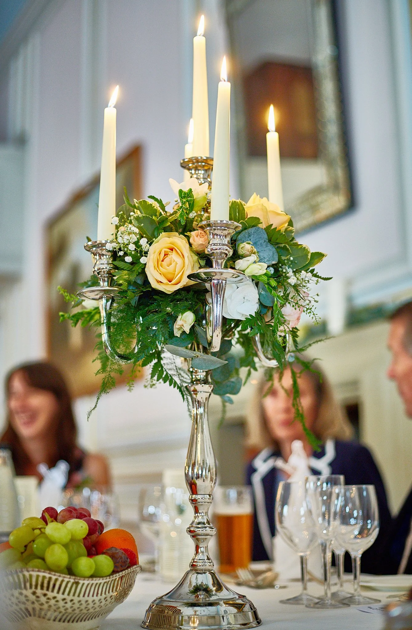 A floral centerpiece with candles on a table at a formal gathering, with people in the background.
