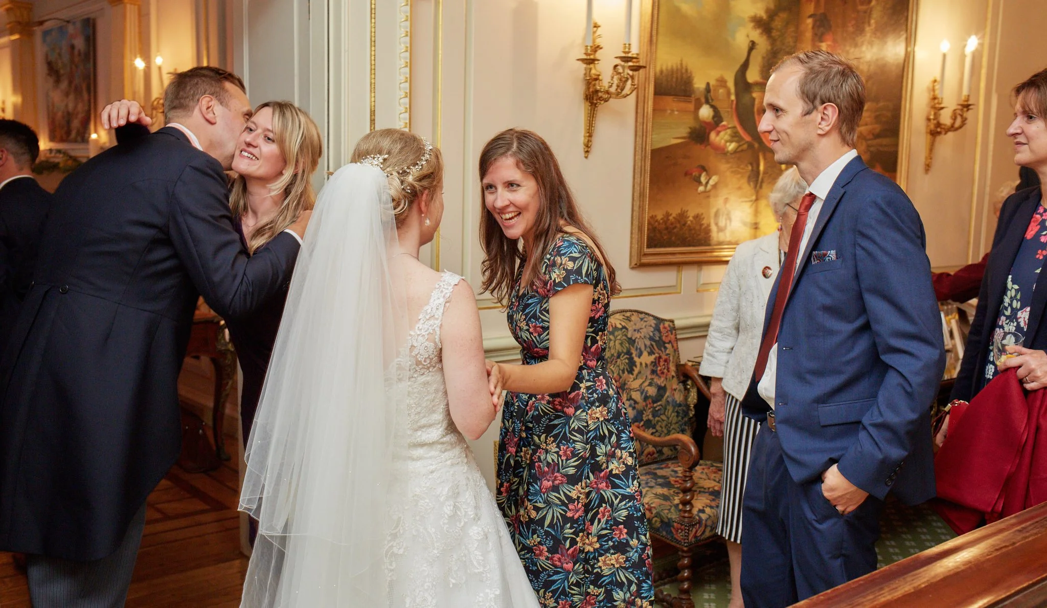 Group of wedding guests socializing indoors, with a bride in white dress and veil, talking with women in floral dresses, and men in suits.