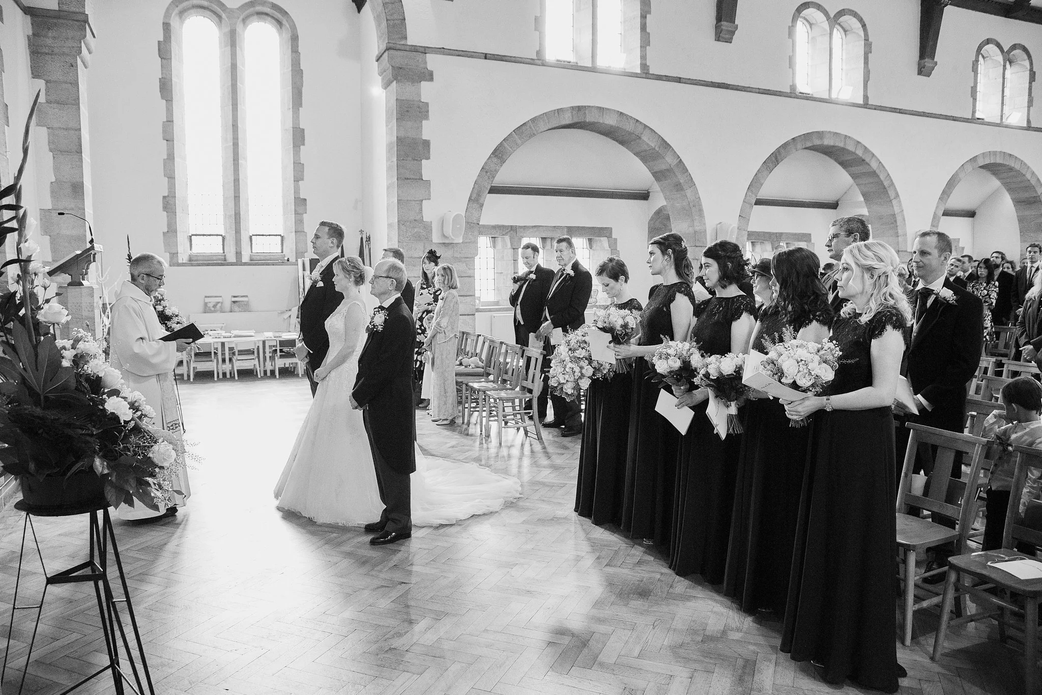 A wedding ceremony inside a church with the bride and groom standing at the altar, surrounded by bridesmaids and groomsmen, all holding flowers and dressed in formal attire. The church has high arched windows and stone walls.