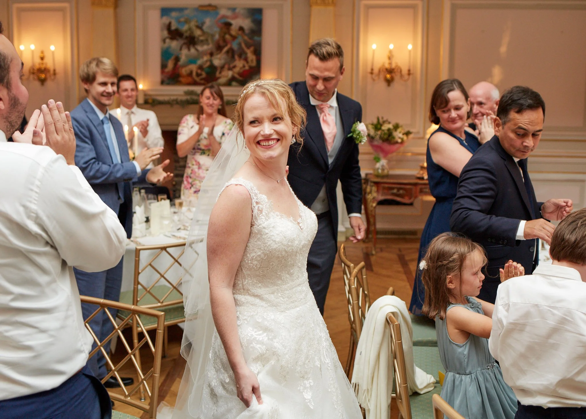 A bride in a white wedding dress smiling at her wedding reception, surrounded by guests clapping and celebrating.