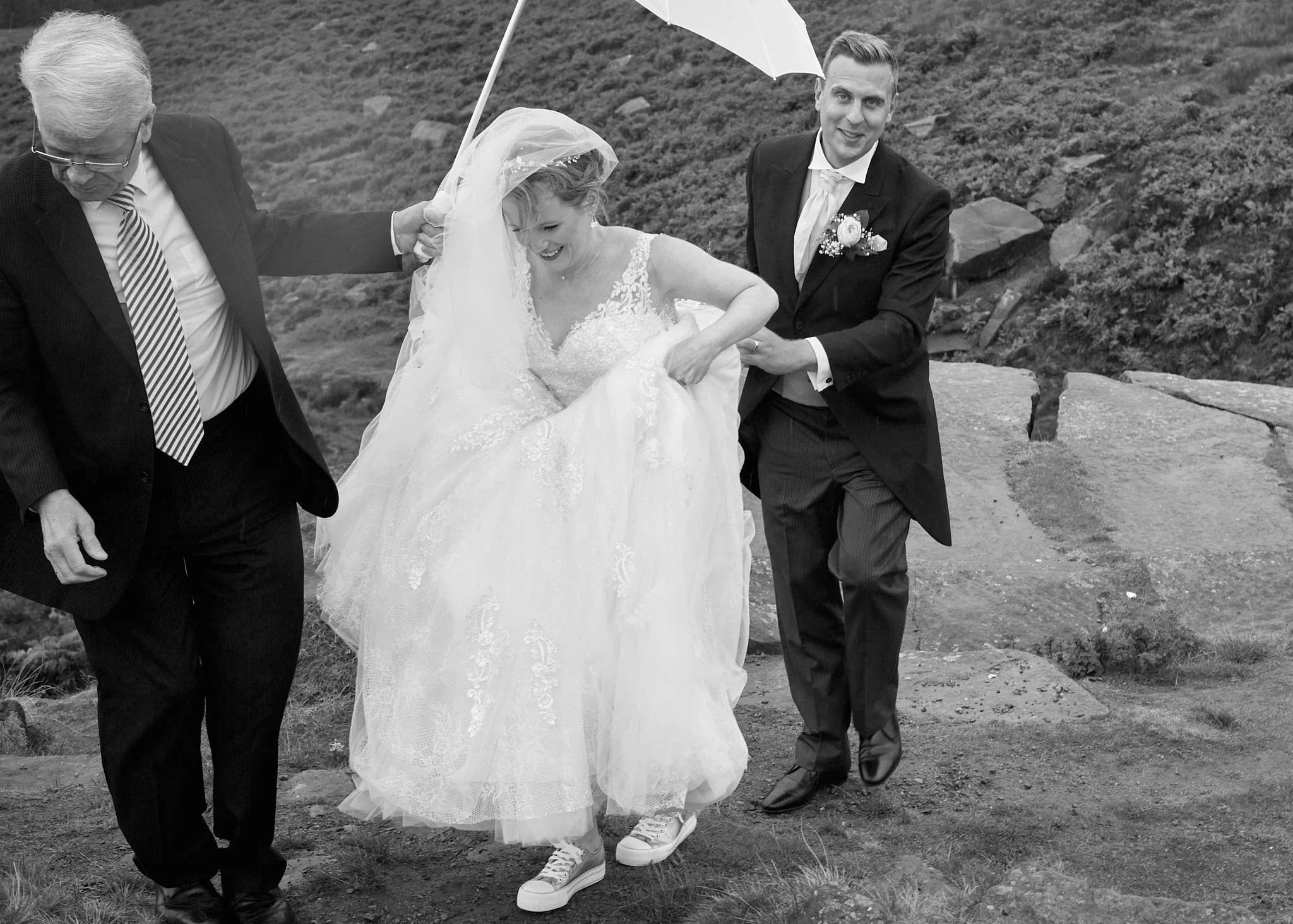 A black and white photo of a wedding ceremony outdoors with three people. A bride in a wedding dress and veil is smiling, with a man in a dark suit holding an umbrella over her. An older man in a pinstripe suit and glasses is holding her hand and wal