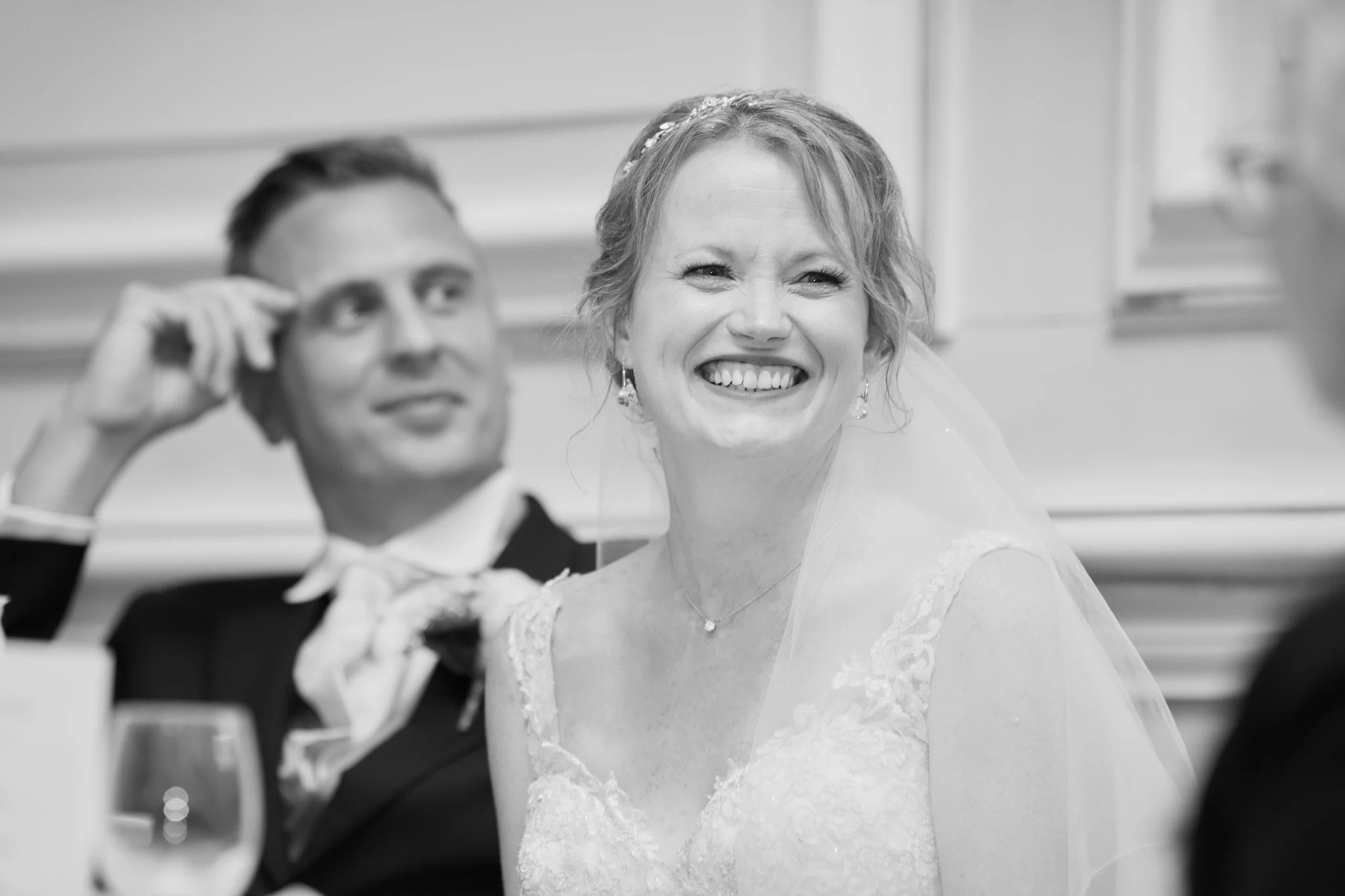 Black-and-white photo of a smiling woman in a wedding dress and a man in a tuxedo, sitting at a table, possibly during a wedding reception.
