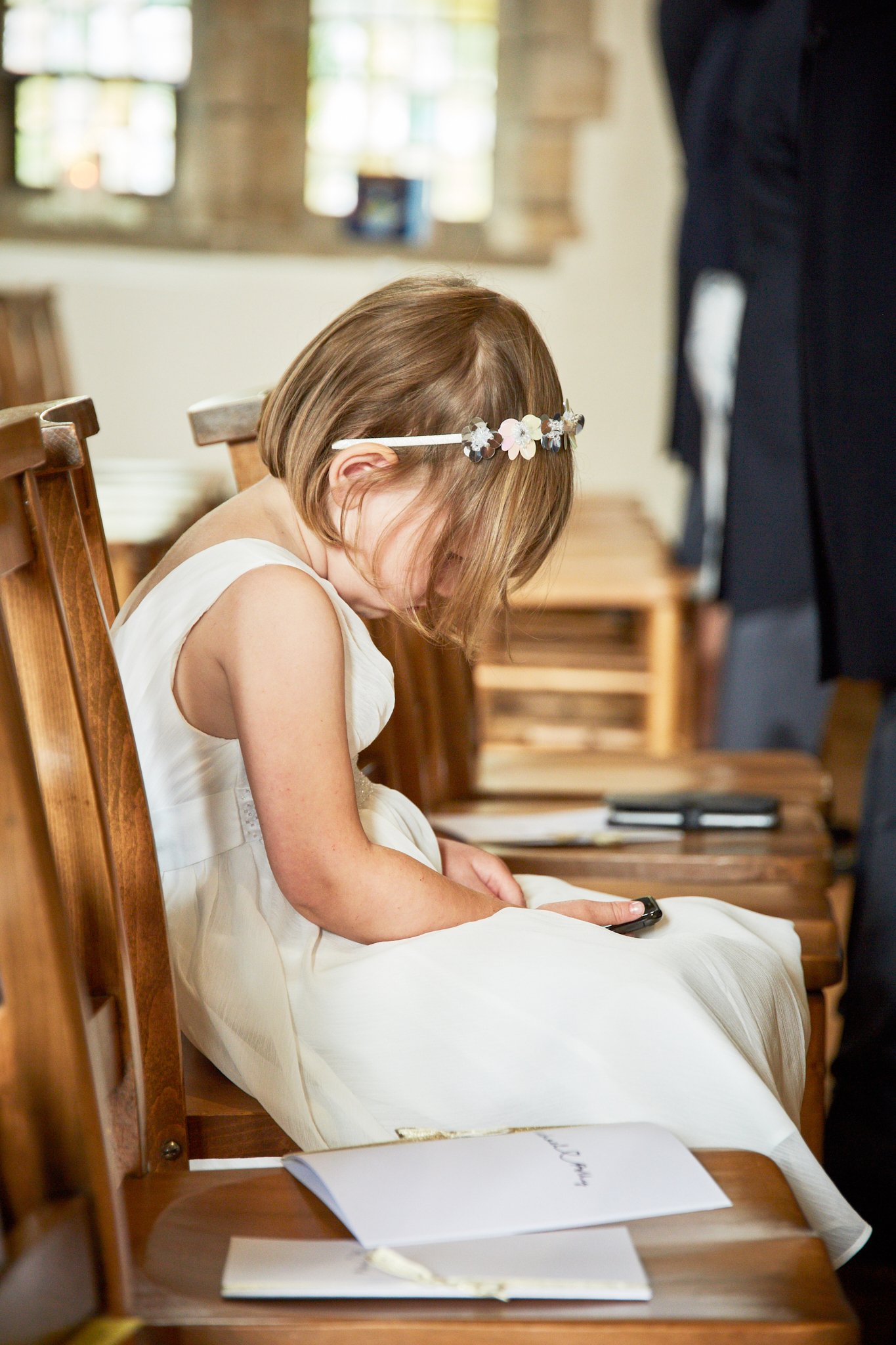 A young girl in a white dress with a floral headband is sitting on a wooden church pew, looking down at a cell phone in her hands during a wedding ceremony.