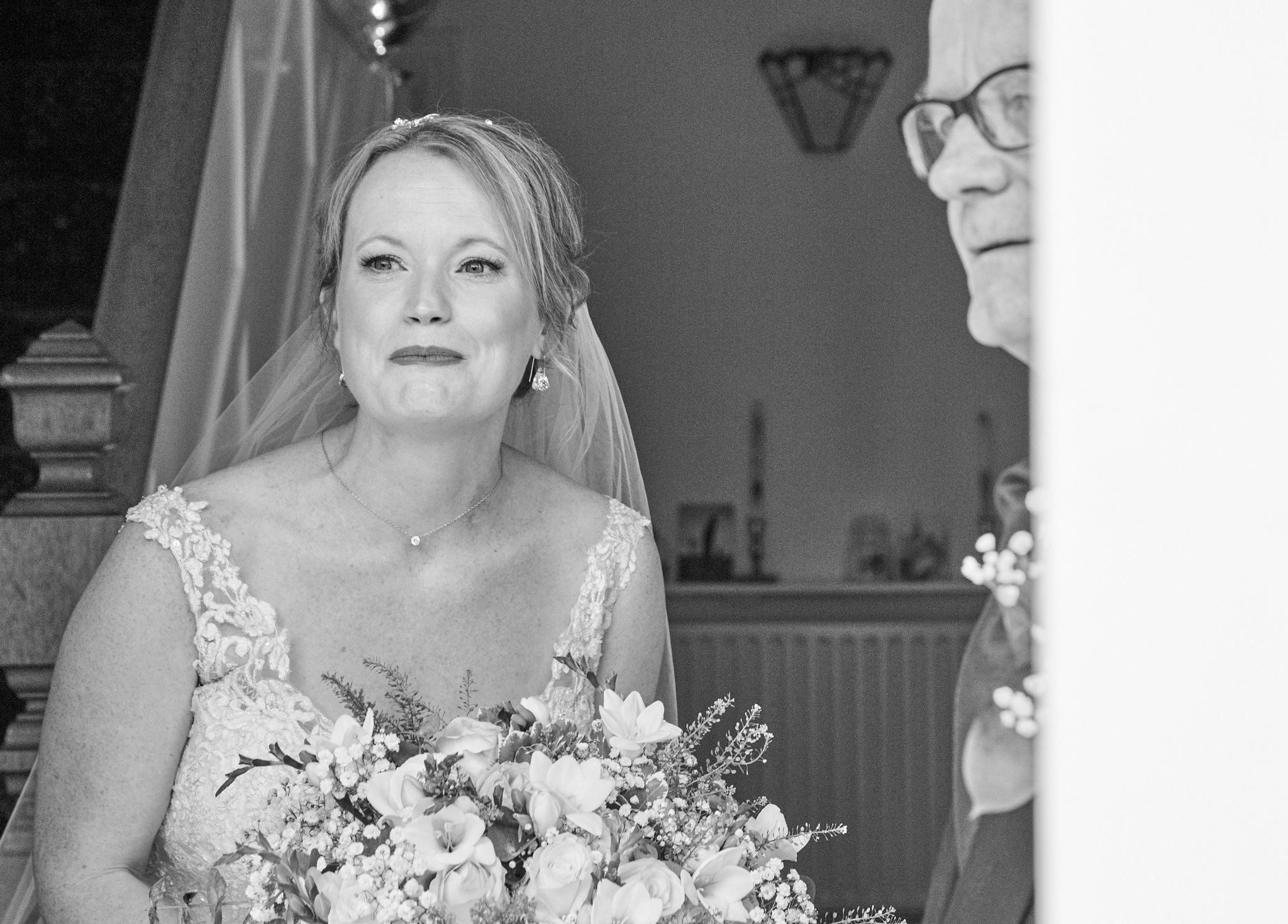 A black and white photo of a bride holding a bouquet of flowers, wearing a lace wedding dress, looking at a man who is partially visible on the right side of the image. The bride has a slight smile on her face.