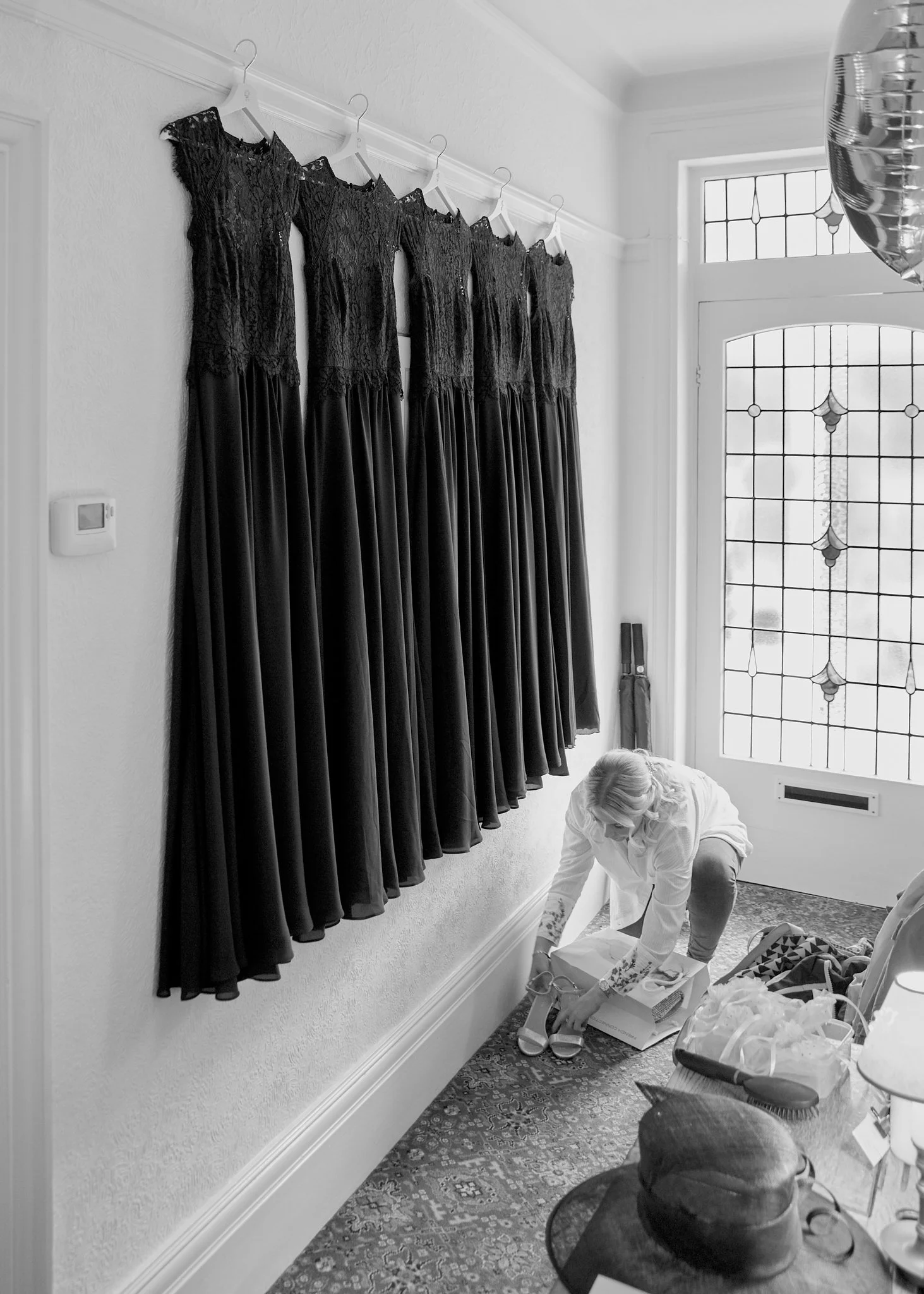 A woman is kneeling on a carpeted floor, unpacking boxes. A row of five black lace dresses is hanging on white hangers on a wall above her. There is a large window with decorative stained glass and a door with a matching window in the background. Var