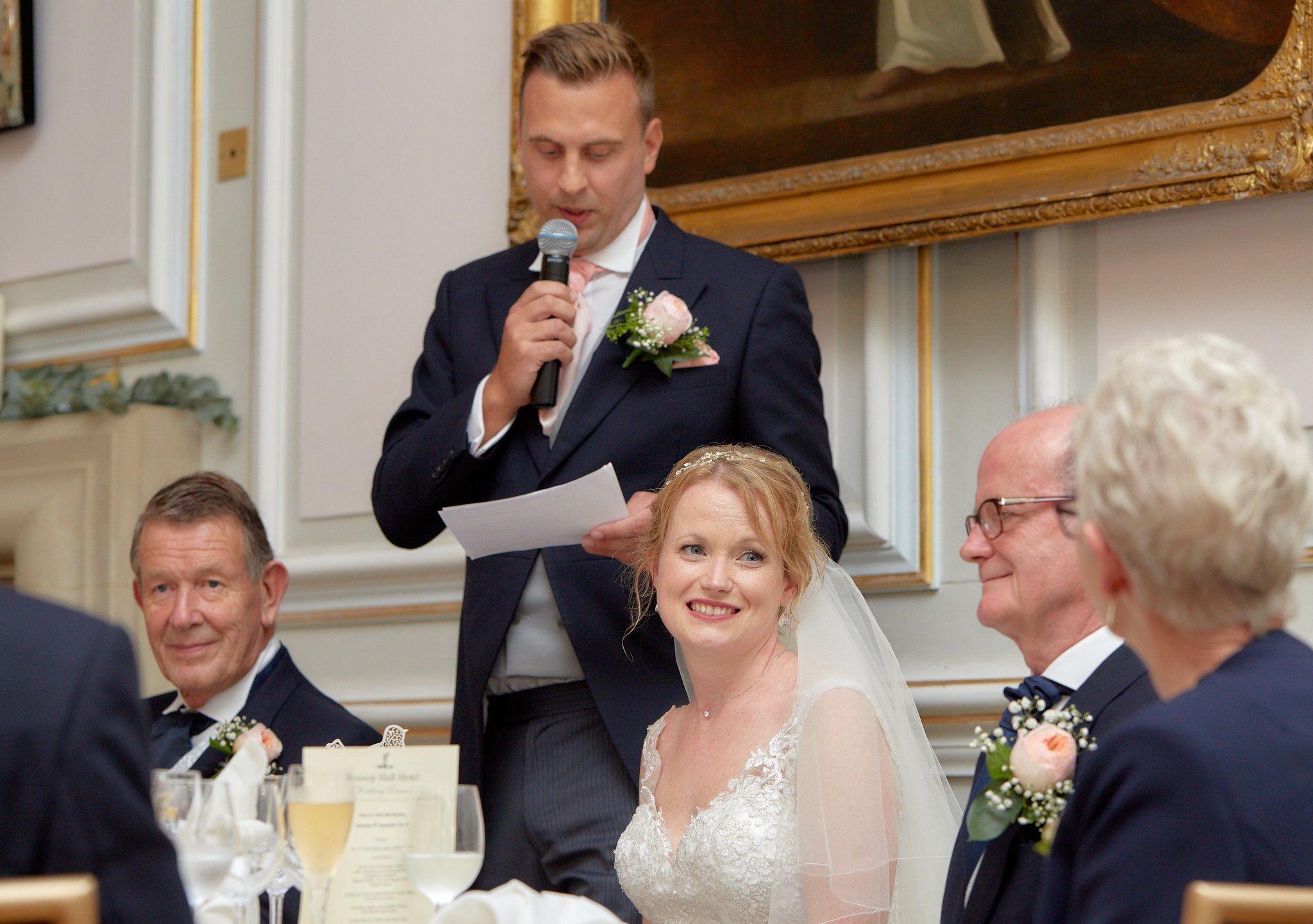 A groom giving a speech at a wedding reception, standing and holding a microphone, with the bride and guests seated at a table.