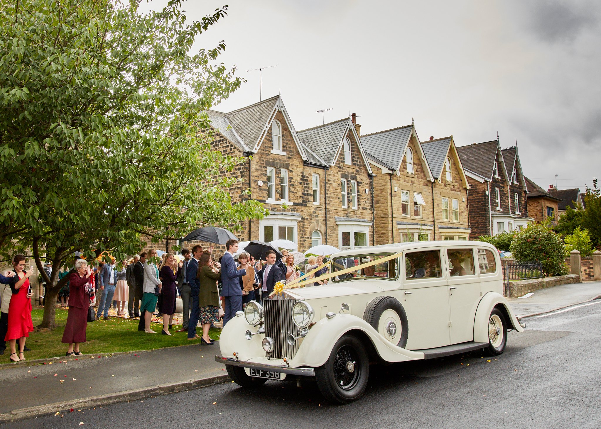 A white vintage car parked on the street during a gathering, with a large group of people dressed in formal attire standing and chatting nearby, in front of a row of old stone houses with gabled roofs and trees, under cloudy skies.