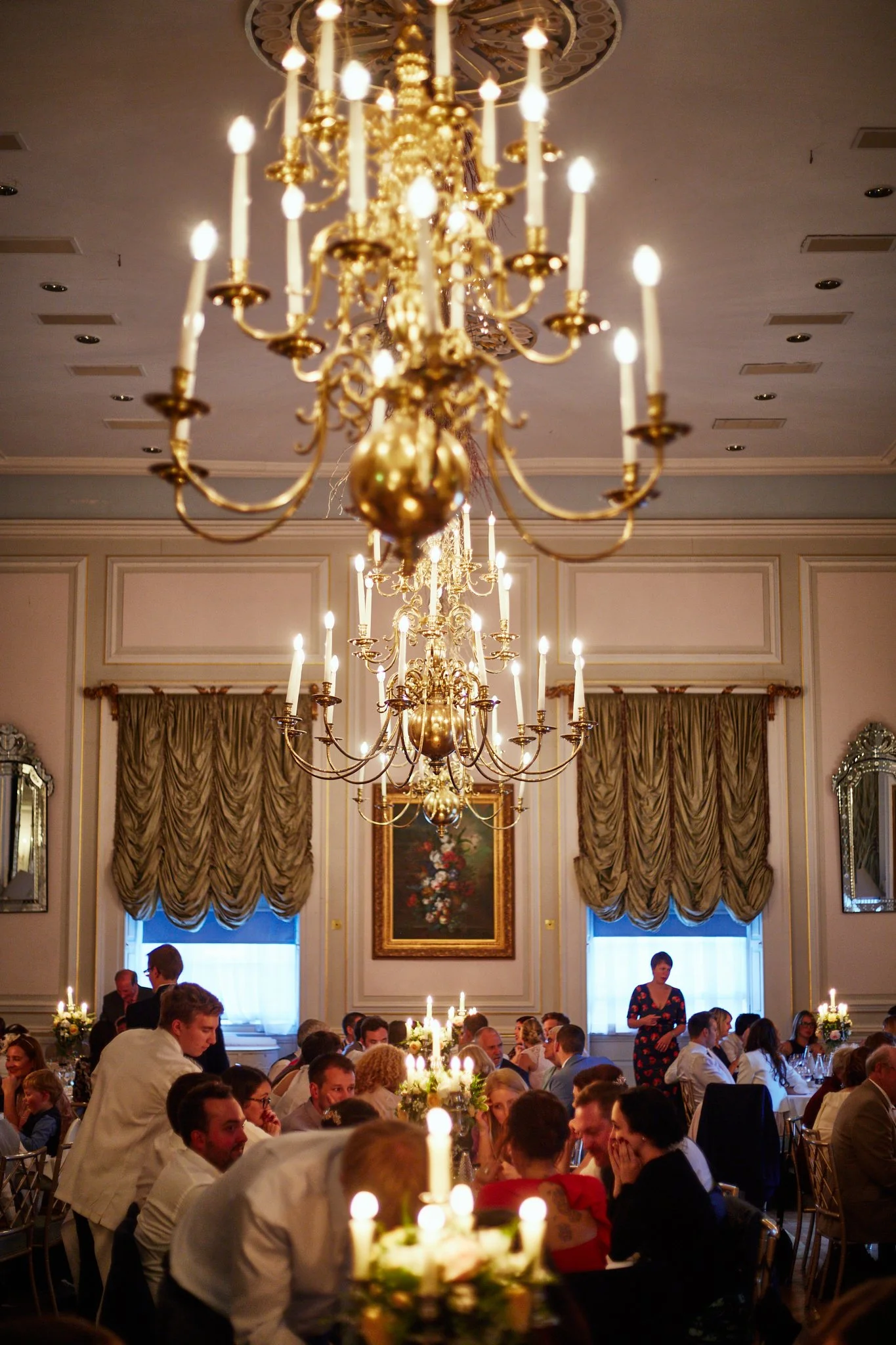 Elegant banquet hall filled with people dining at tables decorated with flowers and candles, large chandeliers hanging from the ceiling, ornate curtains, and framed artwork on the walls.