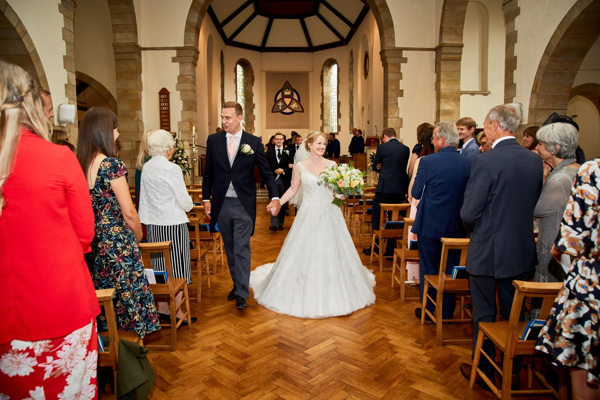 A bride and groom walking down the aisle inside a church, smiling and holding hands, surrounded by seated guests on either side.