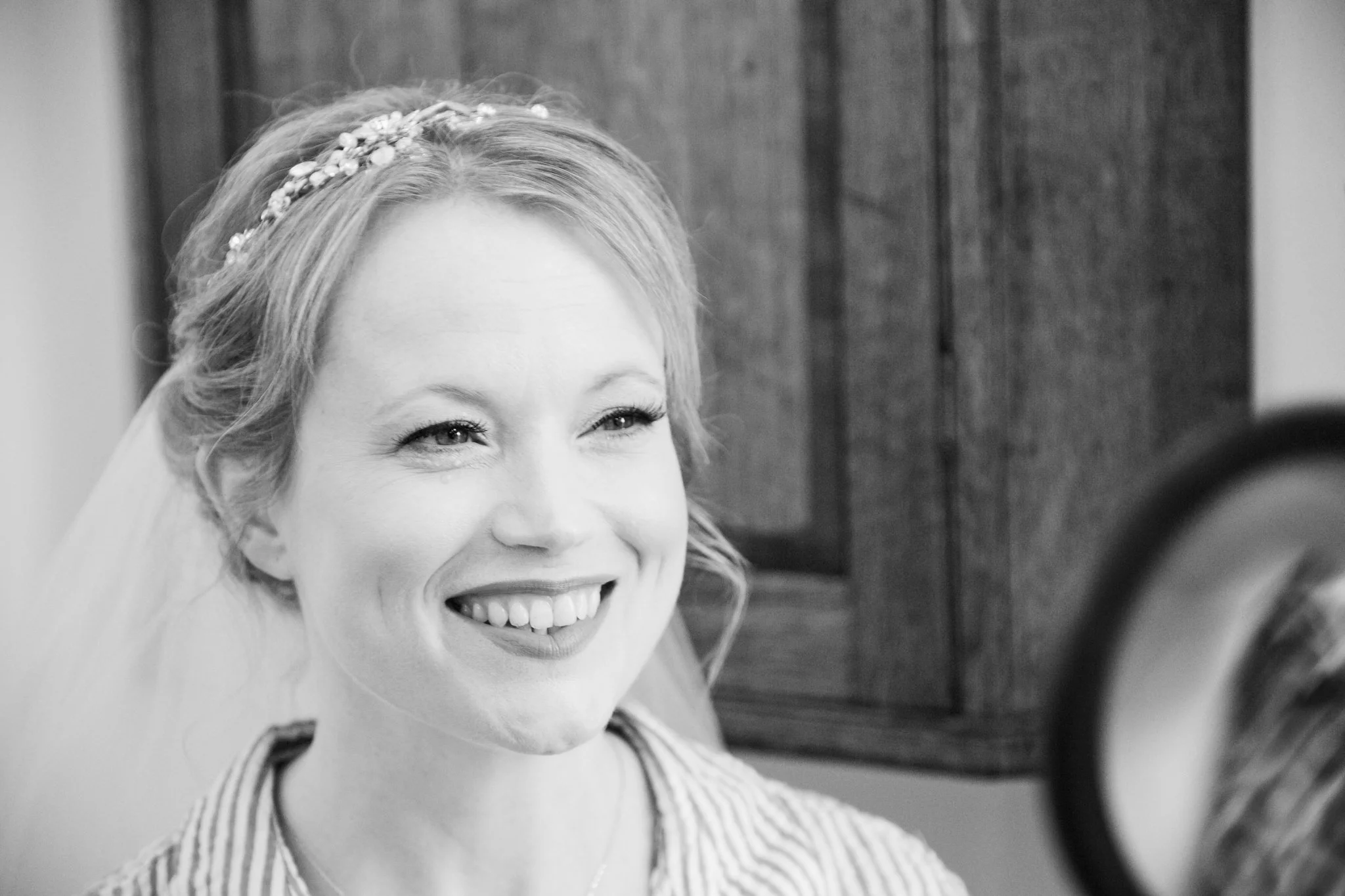 A smiling woman with light hair styled with a decorative headband, wearing a striped top, in a black and white photo, standing in front of a wooden background.