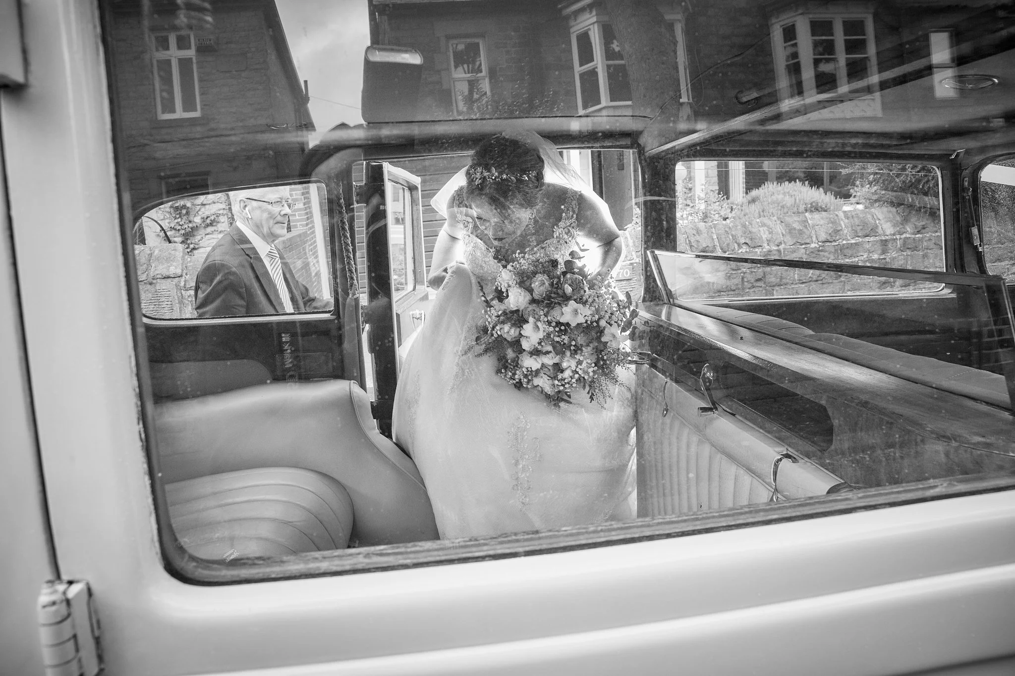A black and white photo of a woman in a wedding dress holding a bouquet of flowers, getting out of a vintage vehicle. An older man in a suit and sunglasses is sitting inside the vehicle, looking at her. The scene is outside on a street with brick hou