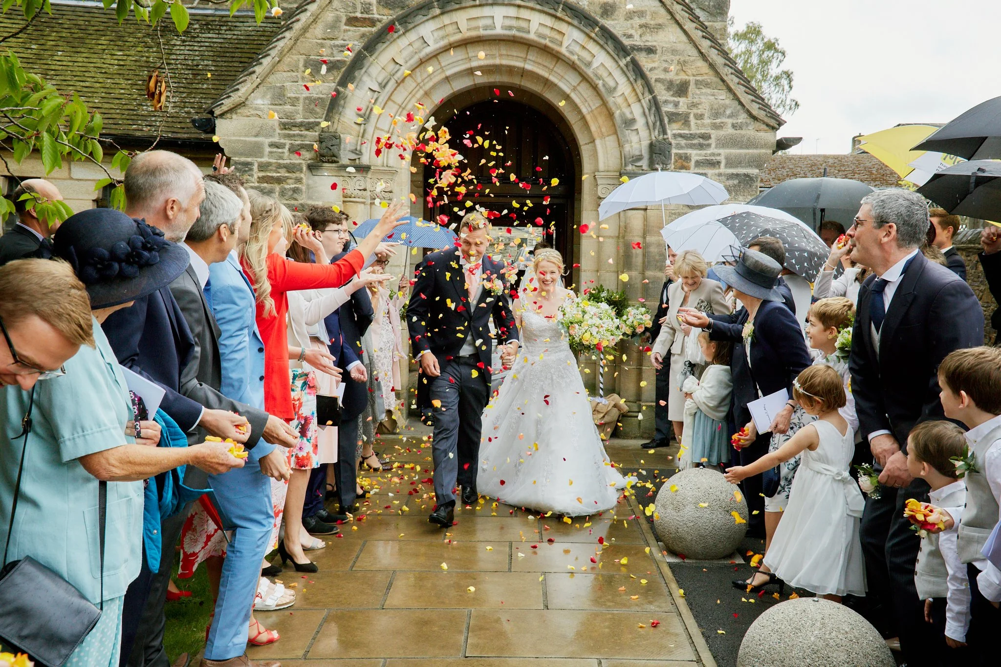 A bride and groom walking out of a church, surrounded by crowd of guests throwing flower petals at them during a wedding celebration.