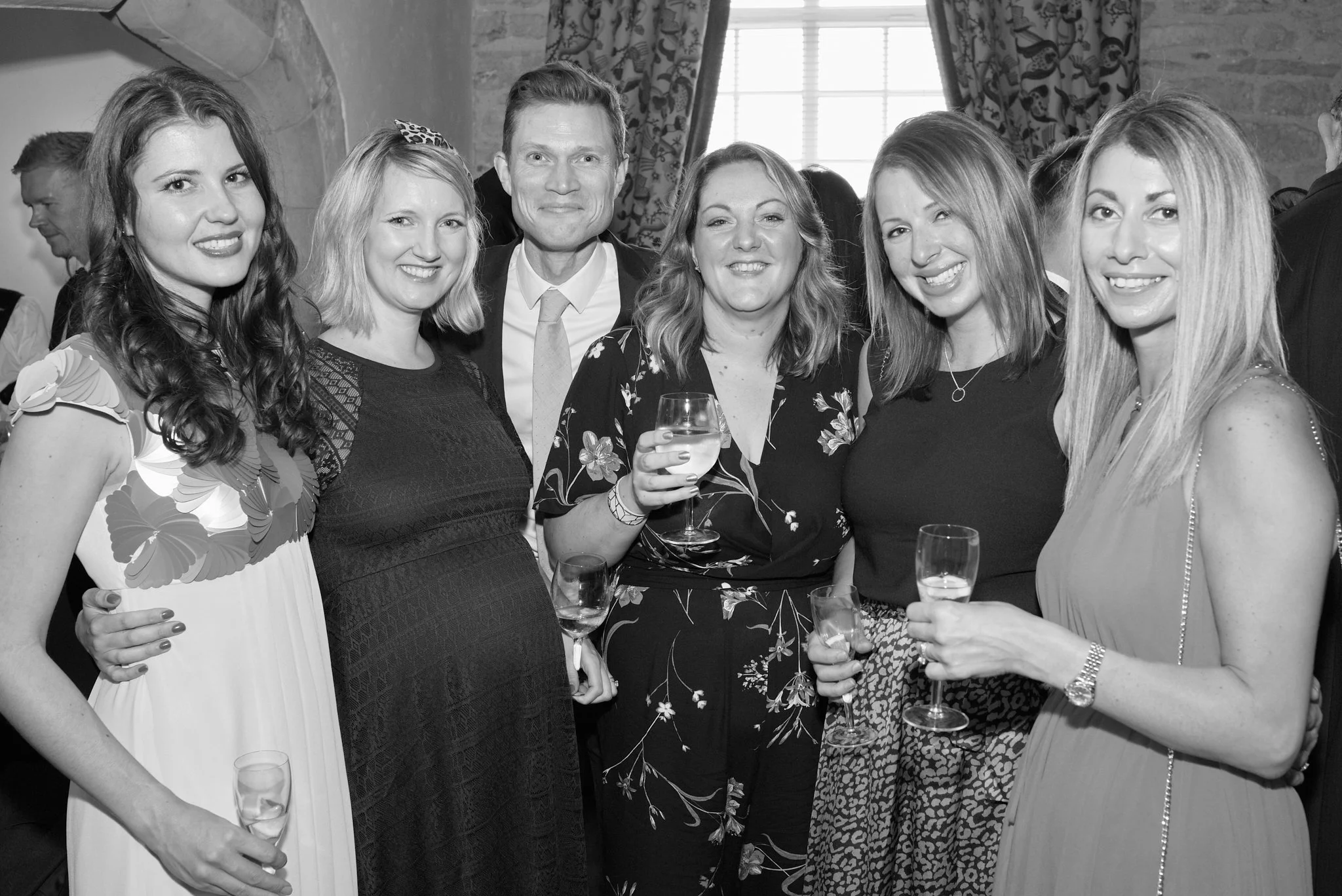 Group of six women and one man standing close together at a social gathering, smiling and holding wine glasses, in a room with a stone wall and large window with curtains.