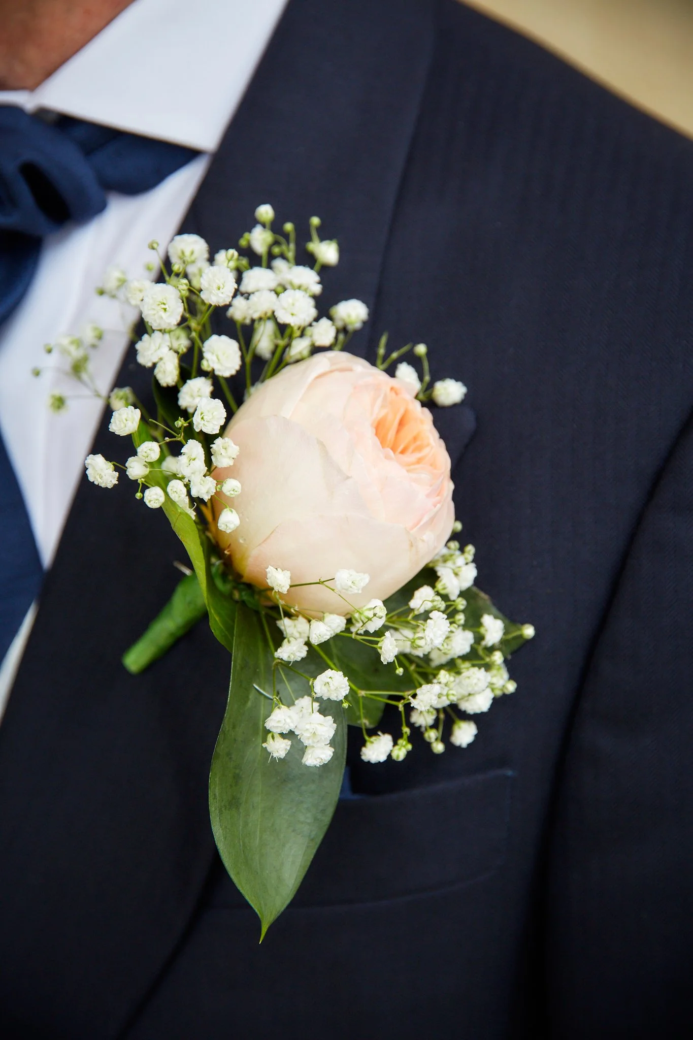 Close-up of a groom wearing a dark suit with a white shirt and a dark bow tie, with a boutonniere made of a pale pink rose and small white flowers pinned to his lapel.