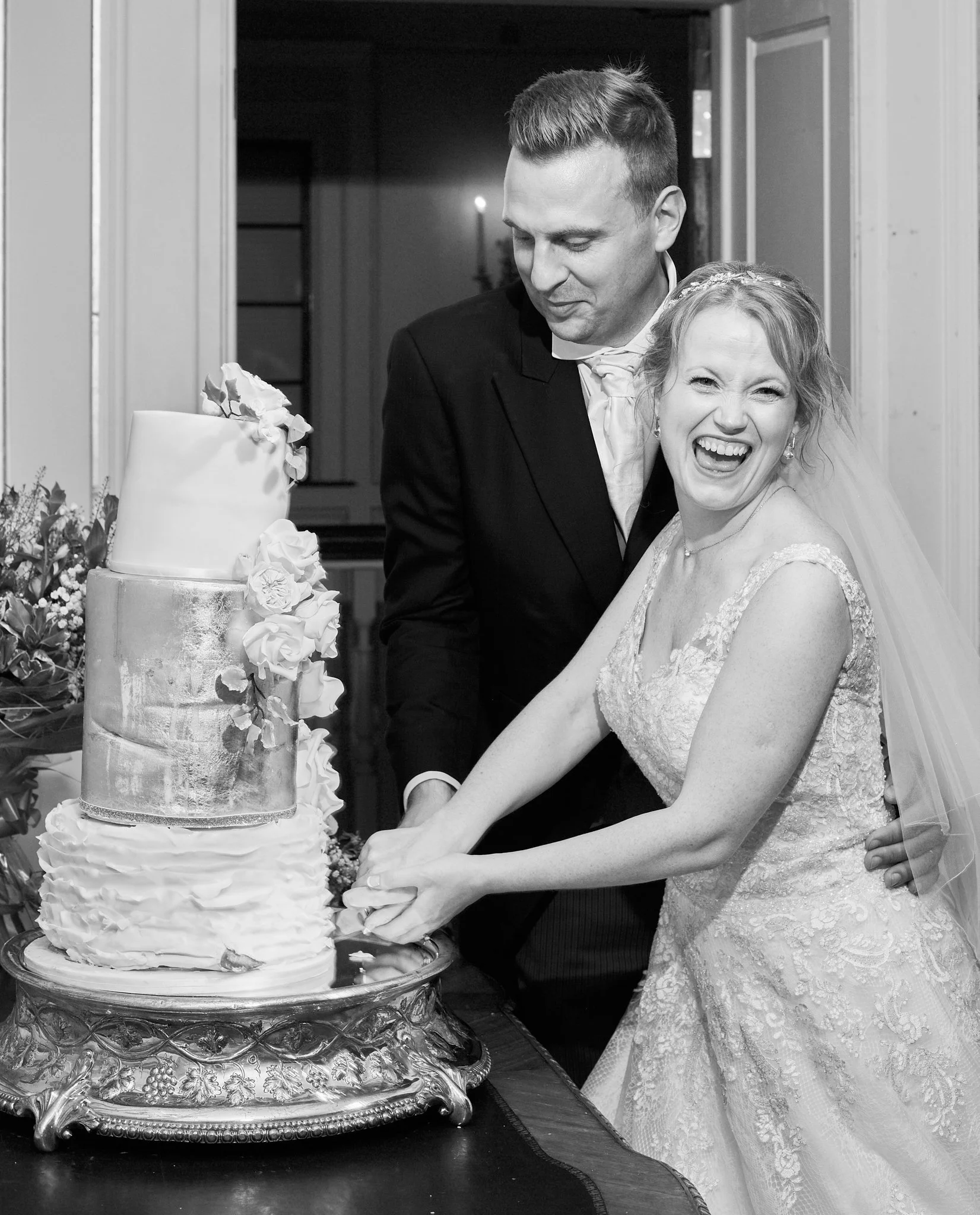 A black and white photo of a bride and groom cutting a wedding cake together at their wedding reception. The bride is smiling and wearing a lace wedding dress with a veil, while the groom is looking at the cake with a slight smile. The wedding cake i
