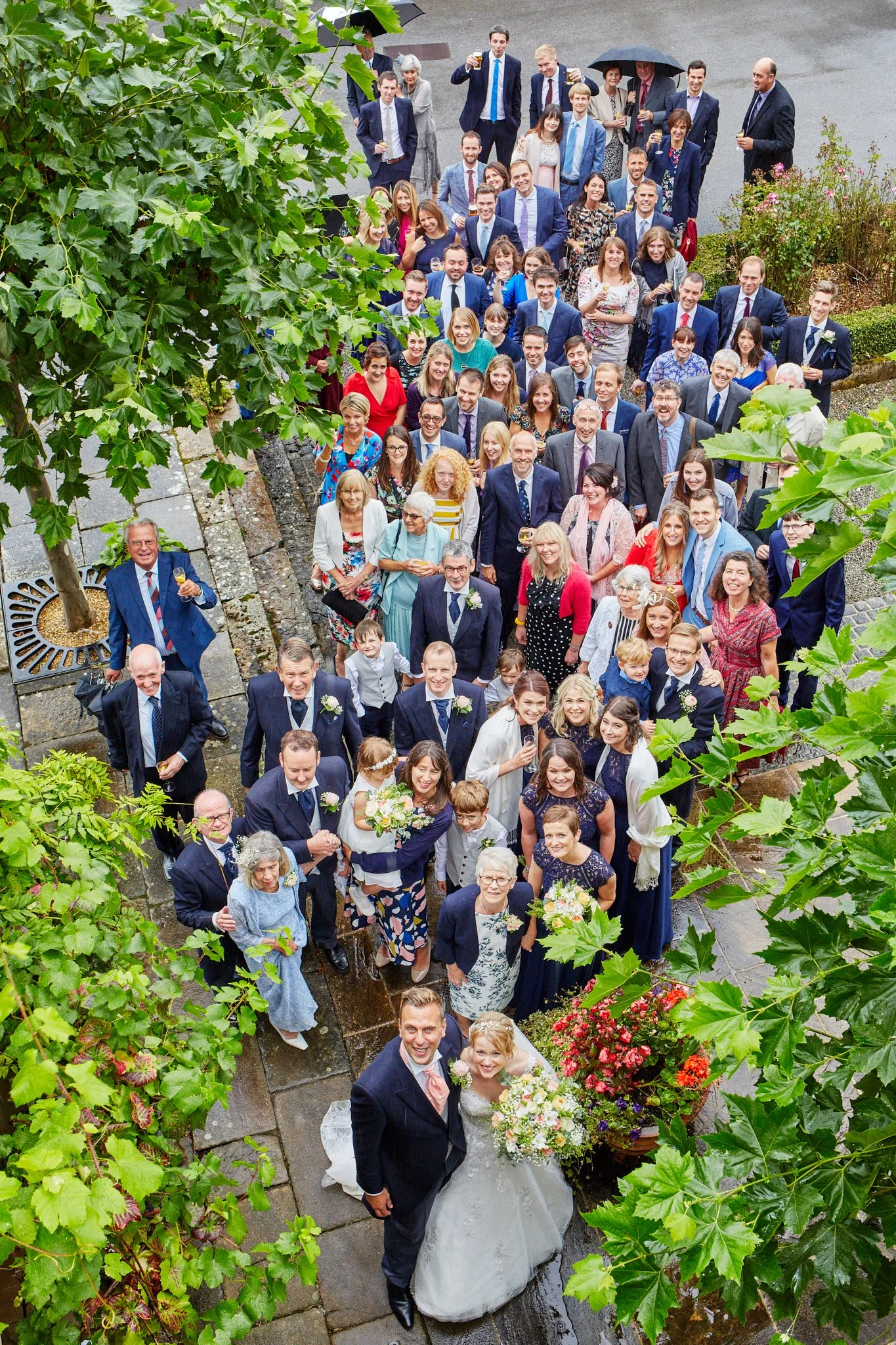 A bride and groom standing together at a wedding reception surrounded by a large group of guests. The guests are dressed in formal attire, holding drinks, and standing outside on a paved courtyard, with greenery and flowers around.