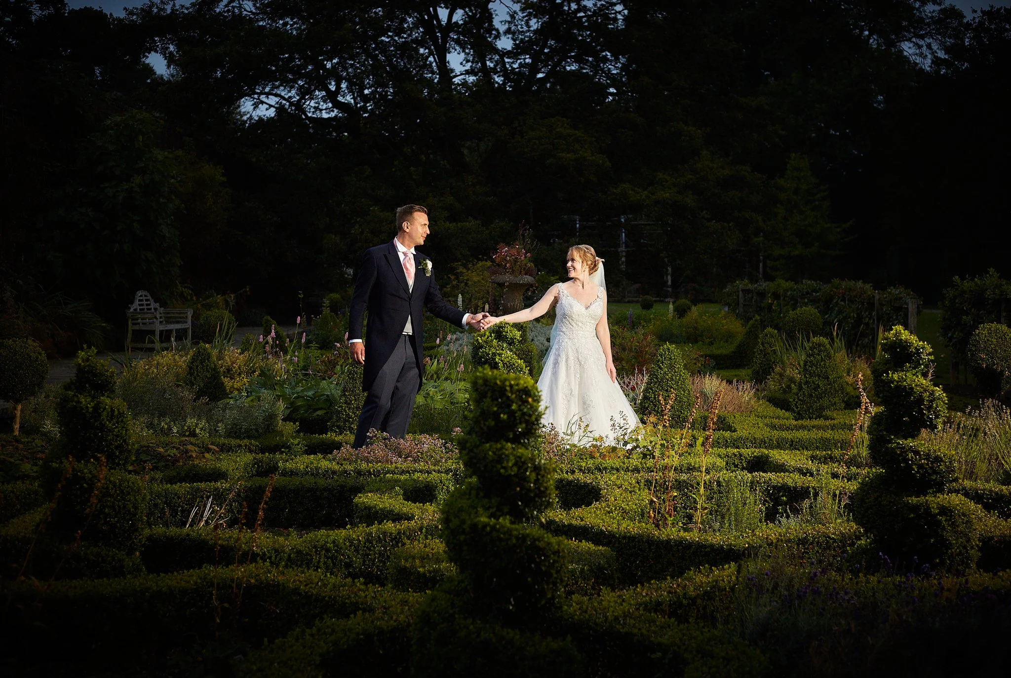 A bride and groom holding hands in a garden during evening sunset, with dark trees and a pathway in the background.