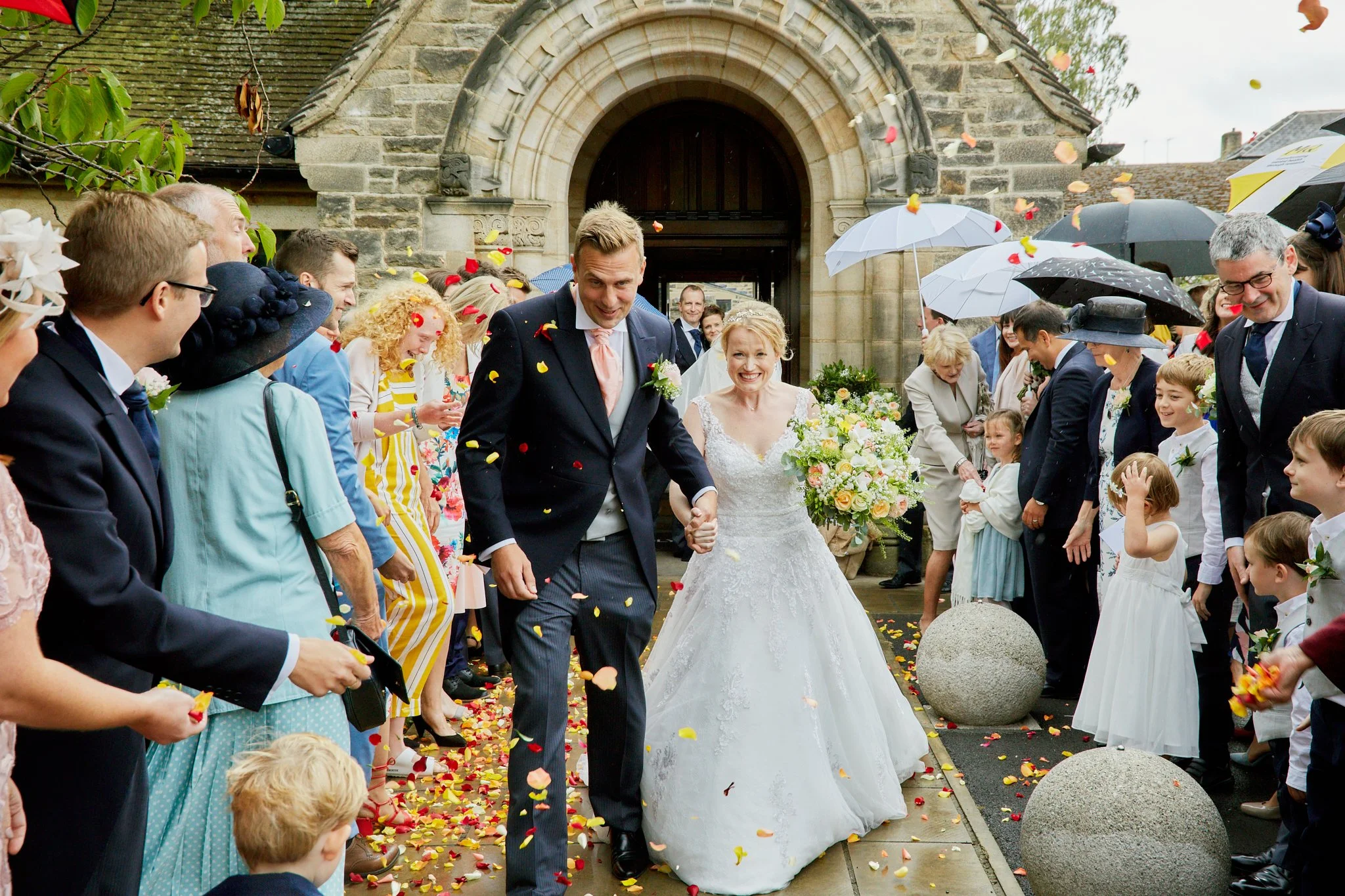 A bride and groom walk out of a church on their wedding day, surrounded by friends and family throwing flower petals and confetti. The bride is holding a bouquet and smiling, while the groom is dressed in a dark suit and tie. Guests are dressed in fo