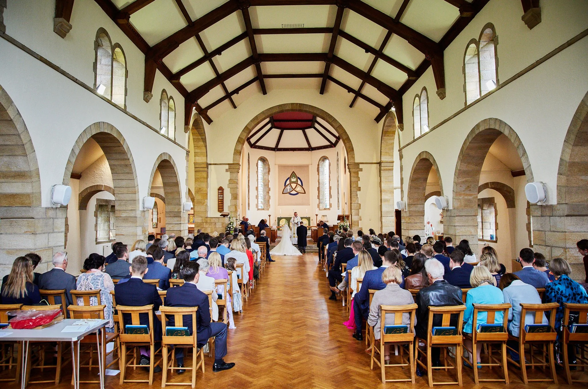 A large congregation inside a church, gathered for a wedding ceremony, with the bride and groom standing at the altar. The church has high vaulted ceilings, stone arches, and tall windows, filled with natural light.